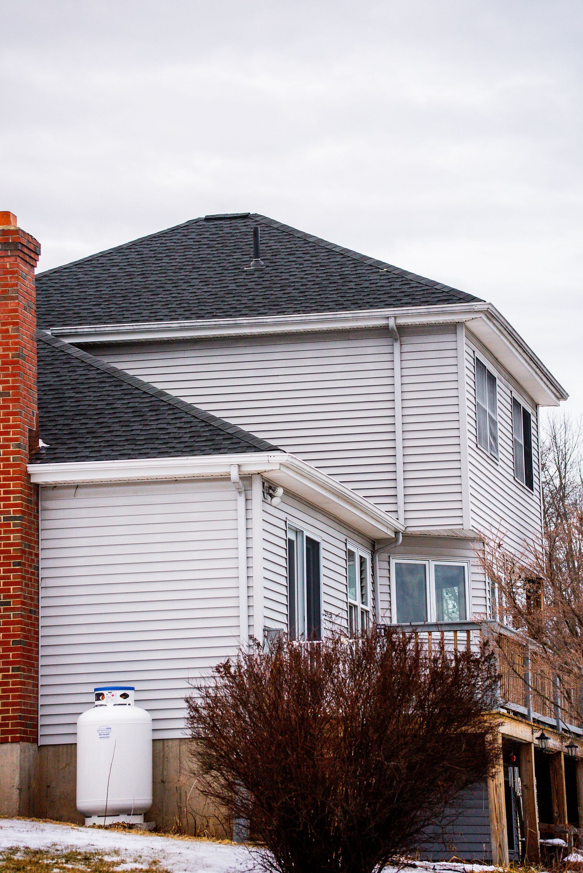 Two-story white house with dark roof and red brick chimney, propane tank, and brown shrubs.