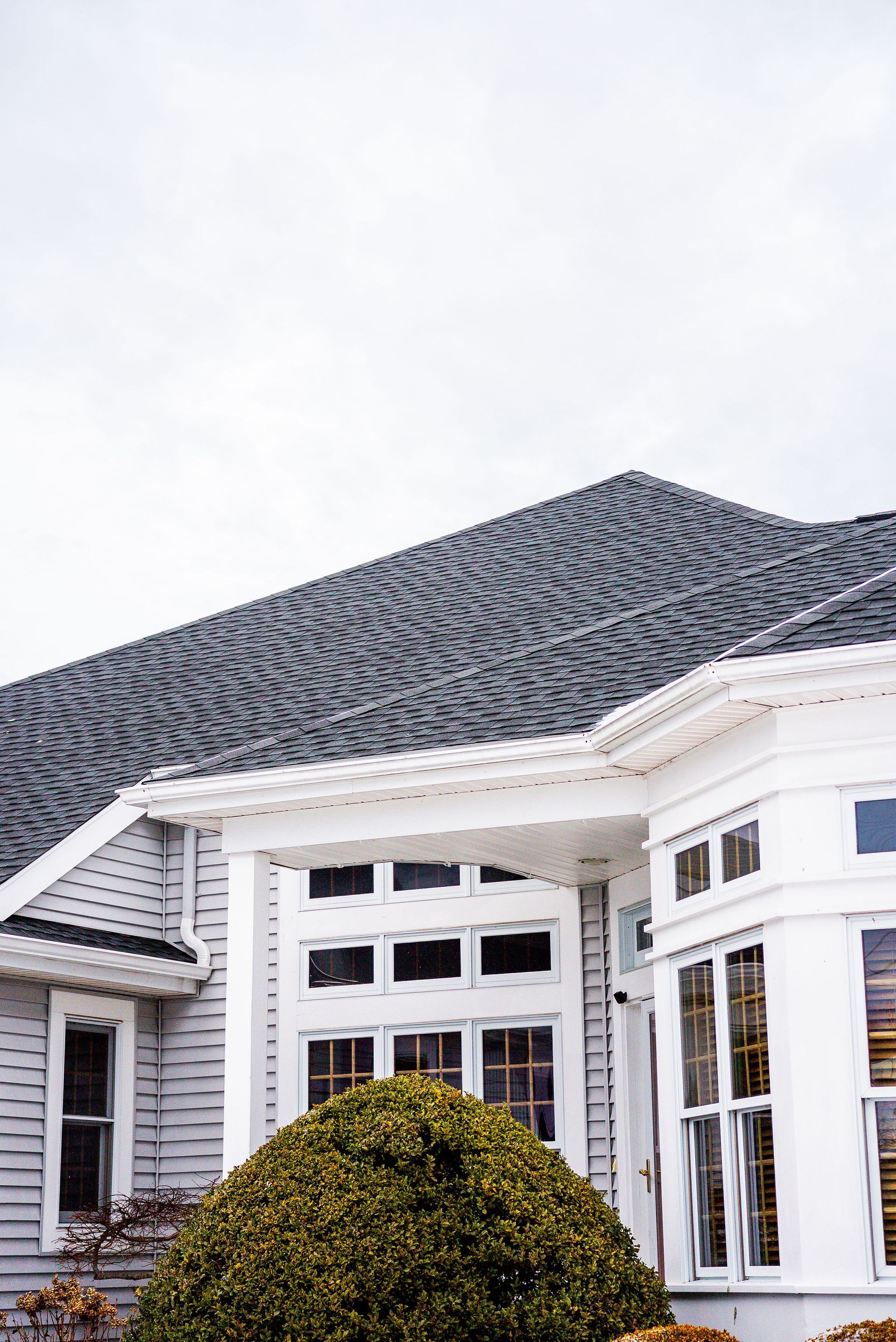 White house with grey roof and windows, with bush in front. Overcast sky.