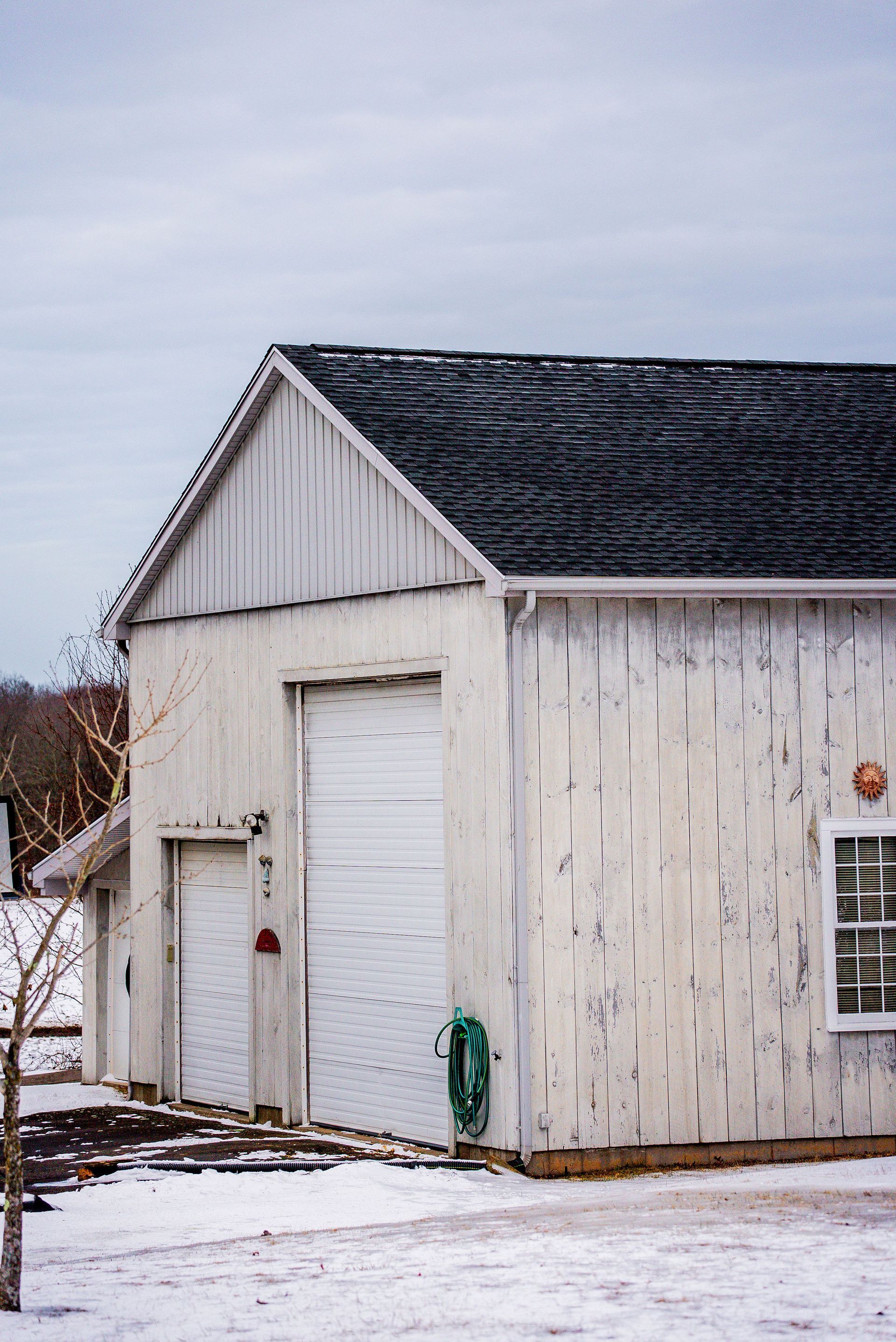 White wooden garage with a closed roll-up door, a window, and a small door, set in a snowy landscape under a cloudy sky.