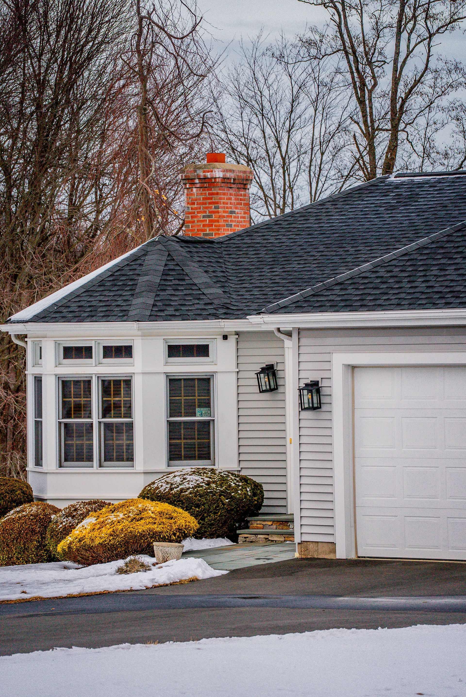 White house with black roof, brick chimney, and garage door. Snow on the ground and bushes.