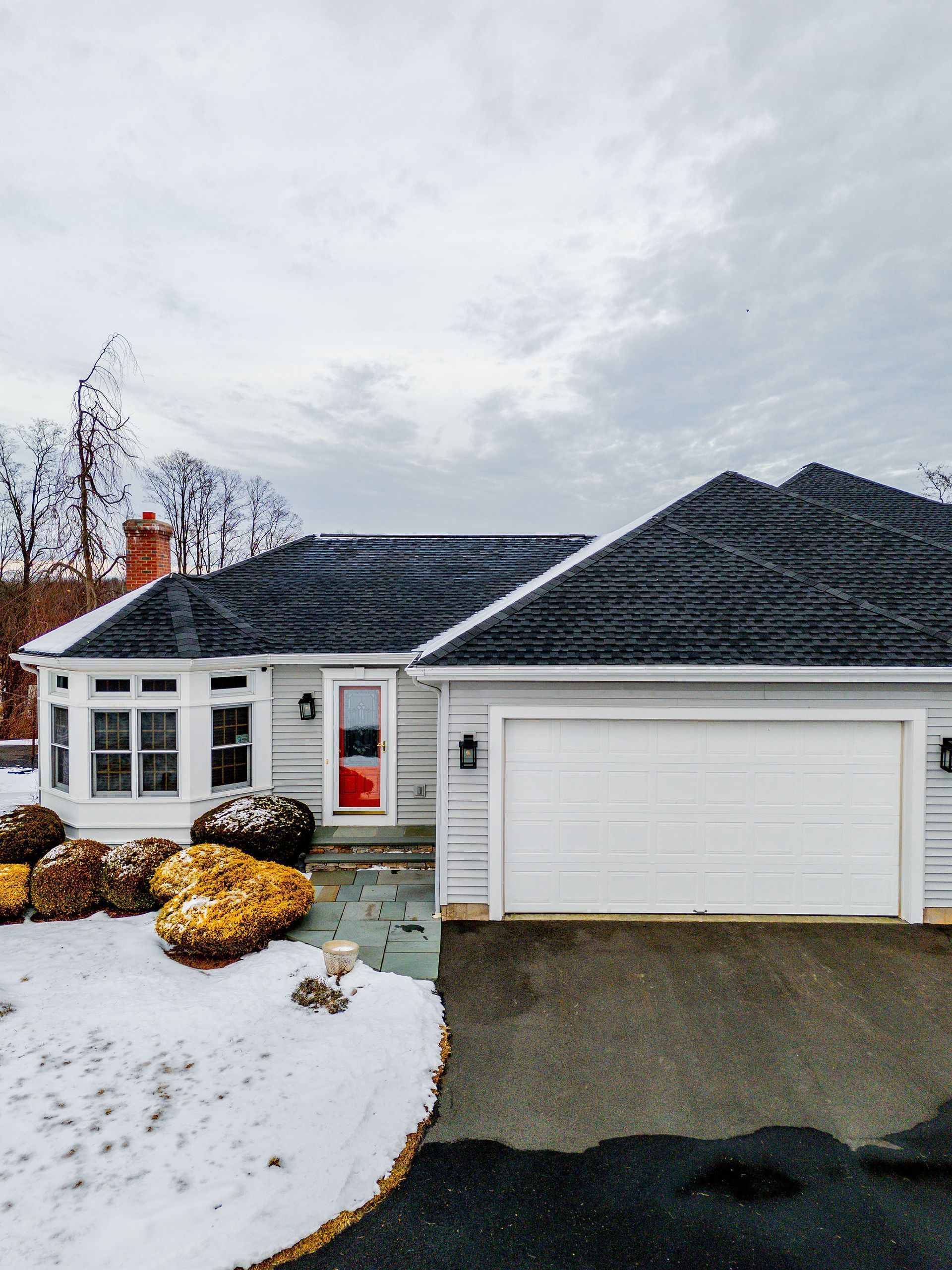 Gray house with dark roof, white garage, red door, snow-covered ground, overcast sky.
