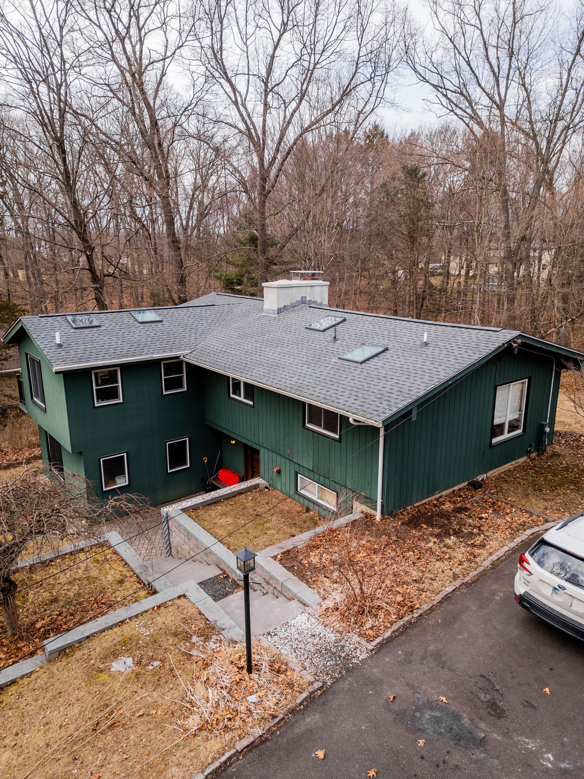 Green house with a dark gray roof, set in a wooded area. Sidewalk leading to red door.