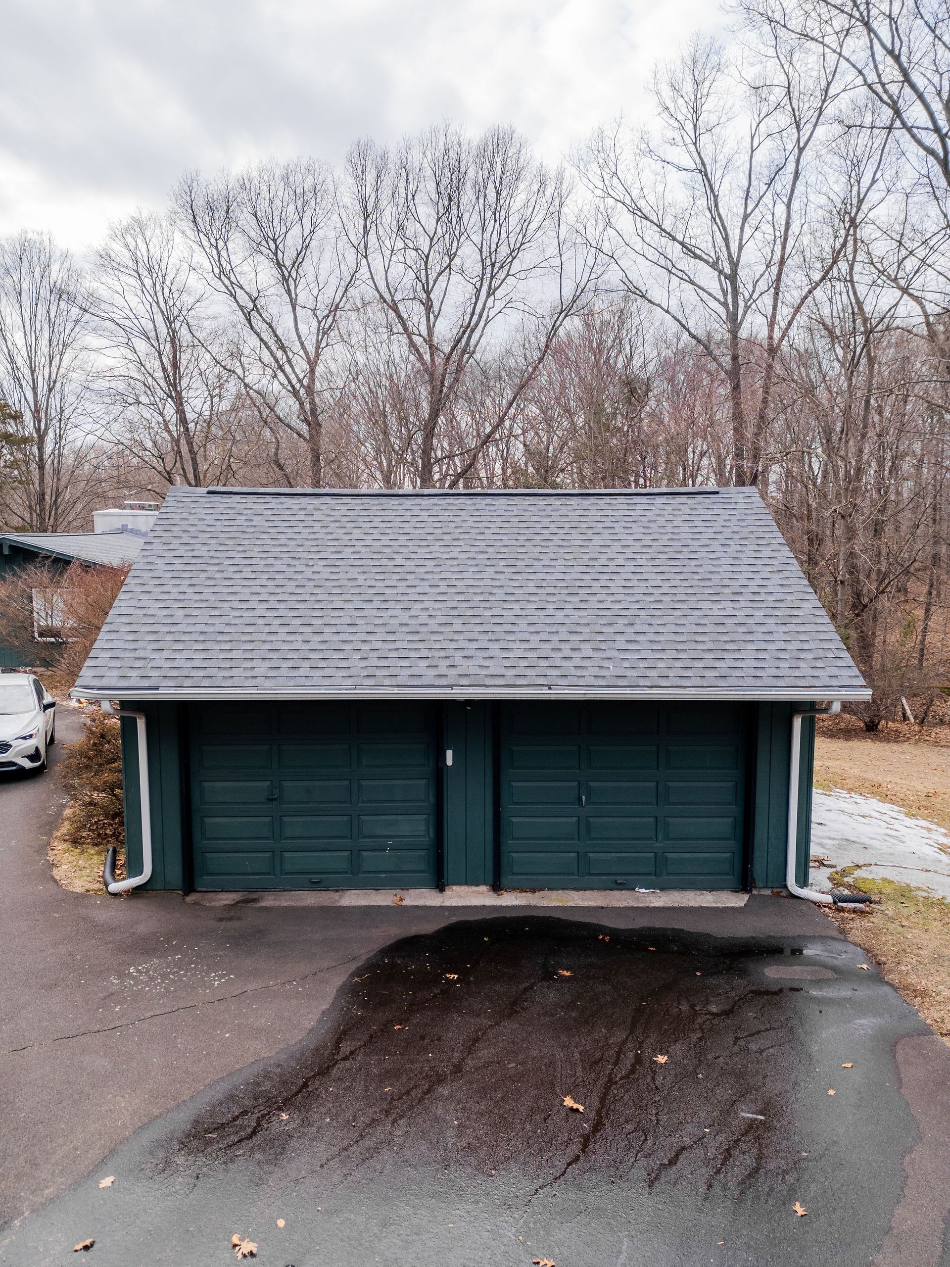 Green double garage with dark roof and driveway in front of bare trees.