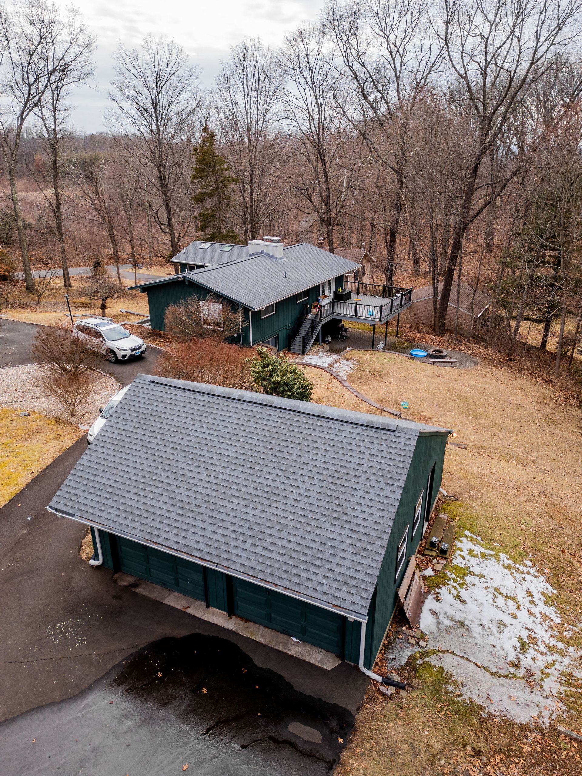 Green house and garage with dark roofs, car parked nearby, trees in the background; winter setting.