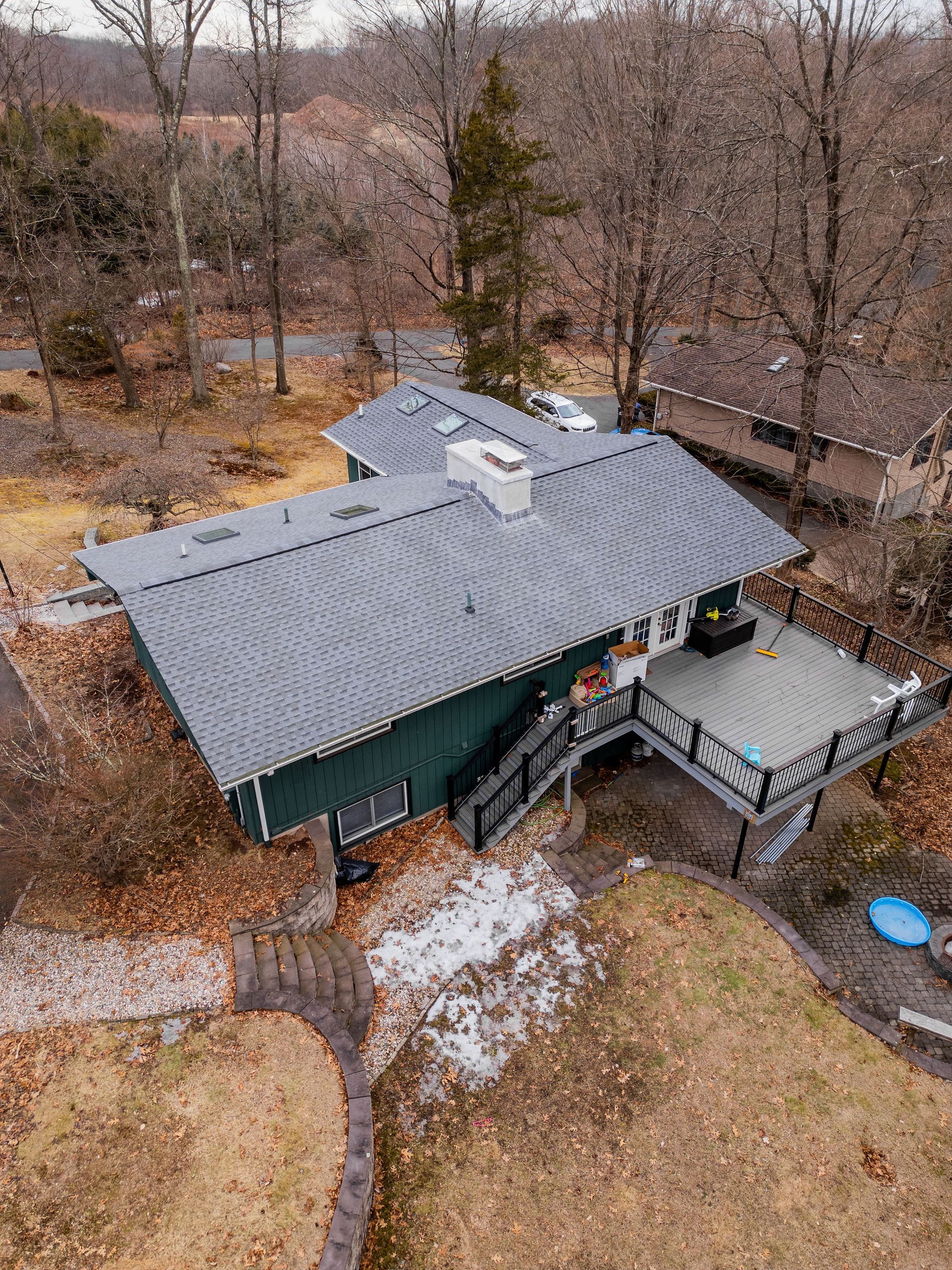 Aerial view of a dark green house with a gray roof and a large wooden deck. Surrounded by trees in a wooded area.