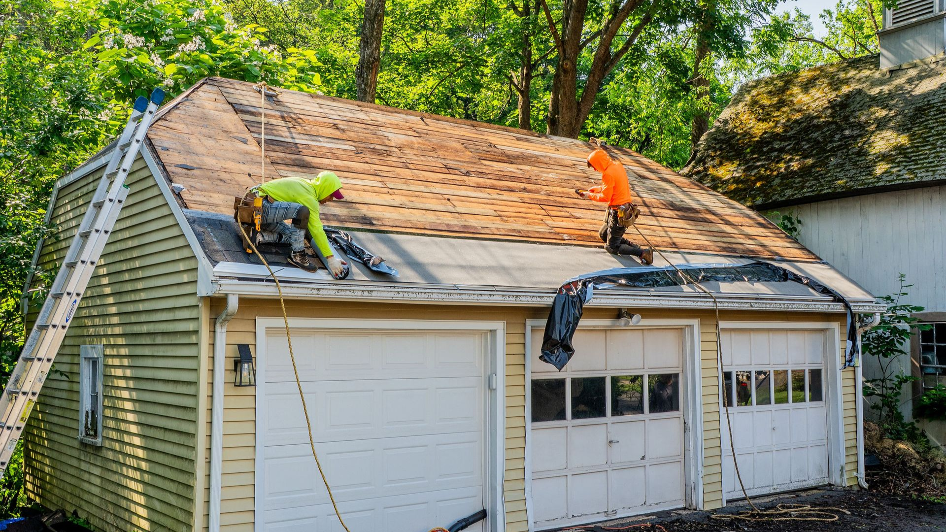 Two roofers working on a garage roof, removing shingles. Yellow siding, white garage doors, trees in the background.