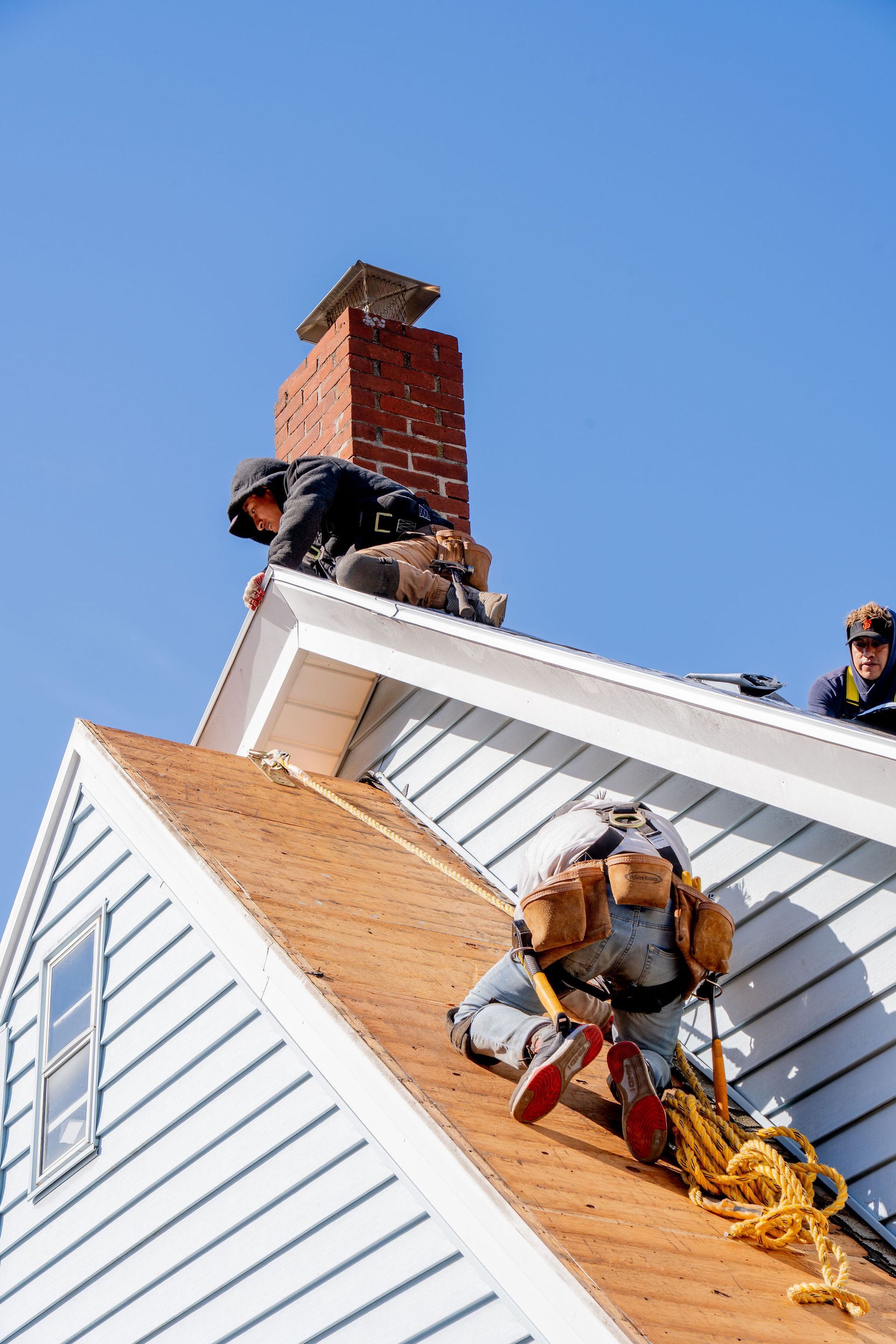 Roofers work on a shingled roof, near a brick chimney, under a blue sky.