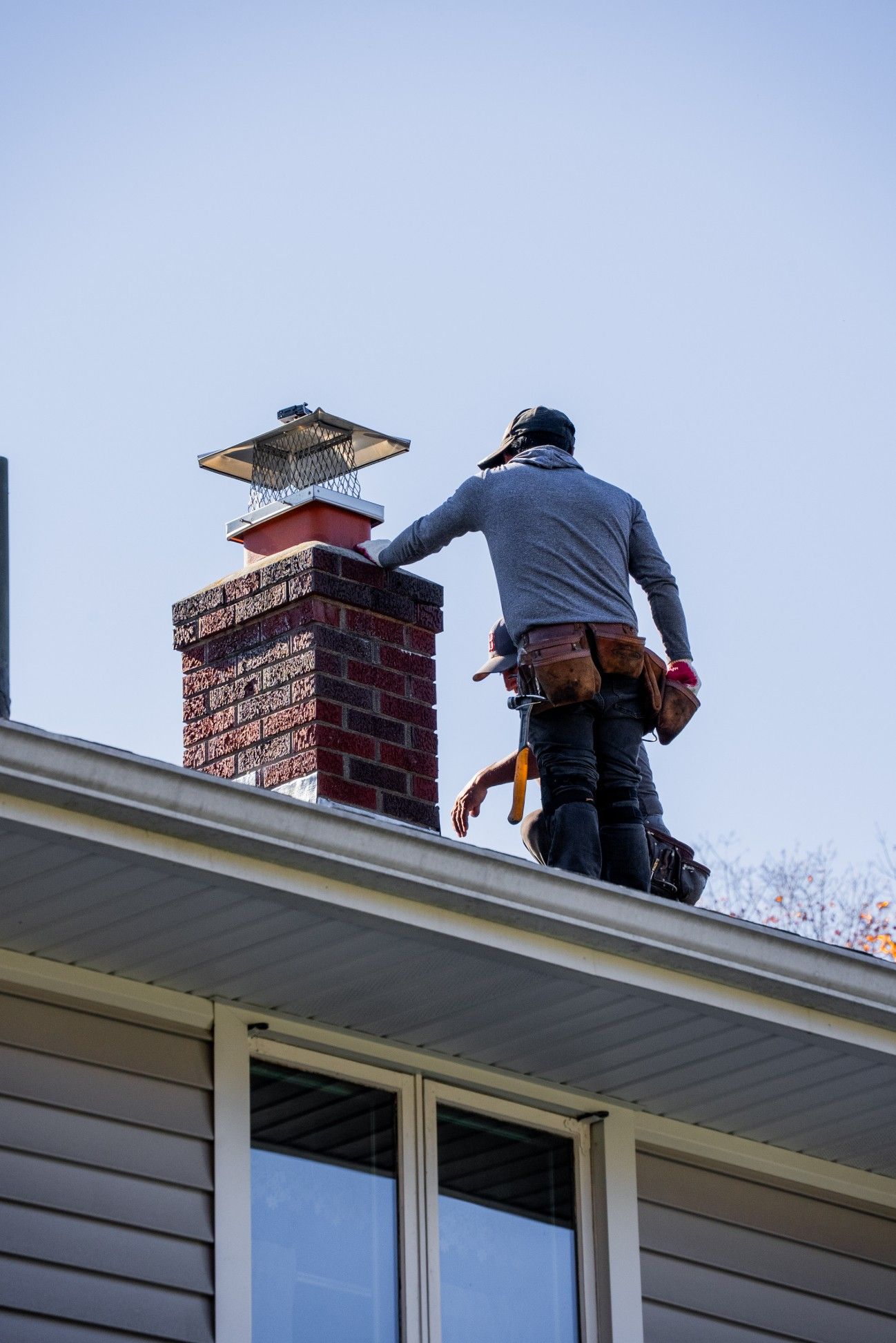 A person wearing a grey hoodie and tool belt stands on a sloped roof next to a brick chimney with a metal cap.