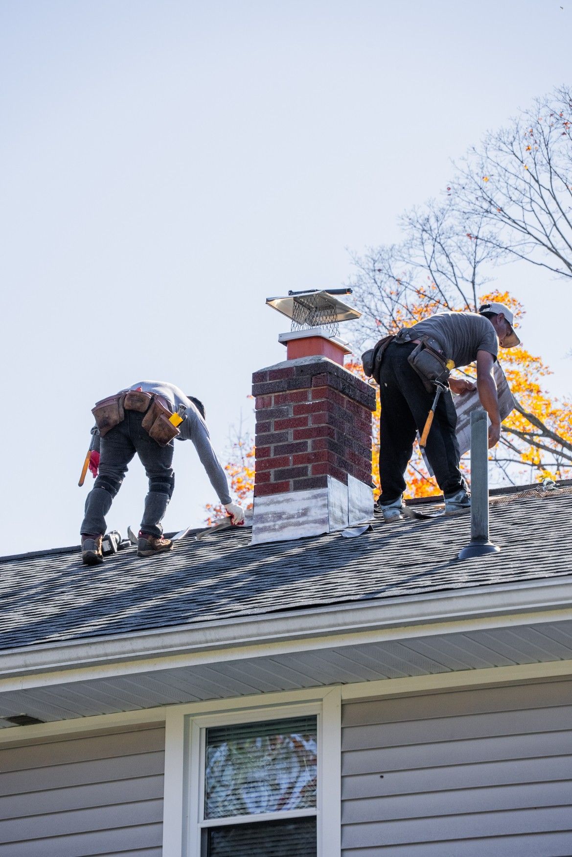 Two workers in tool belts stand on a shingled residential roof, working near a brick chimney.