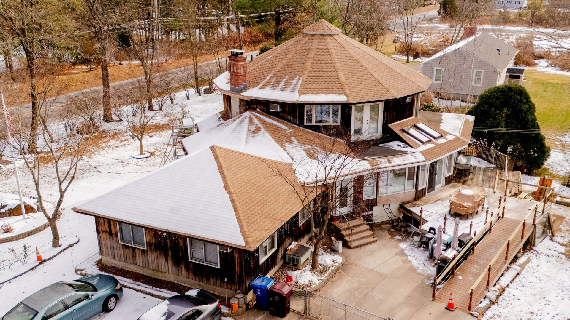 Two-story brown house with a round roof and attached deck, covered in snow, with bare trees and a car in the driveway.