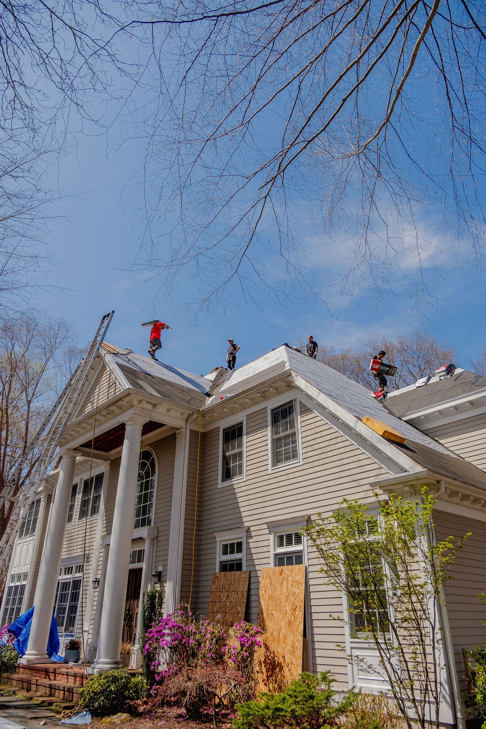 Roofers work on a large, light-colored house with columns under a bright blue sky.