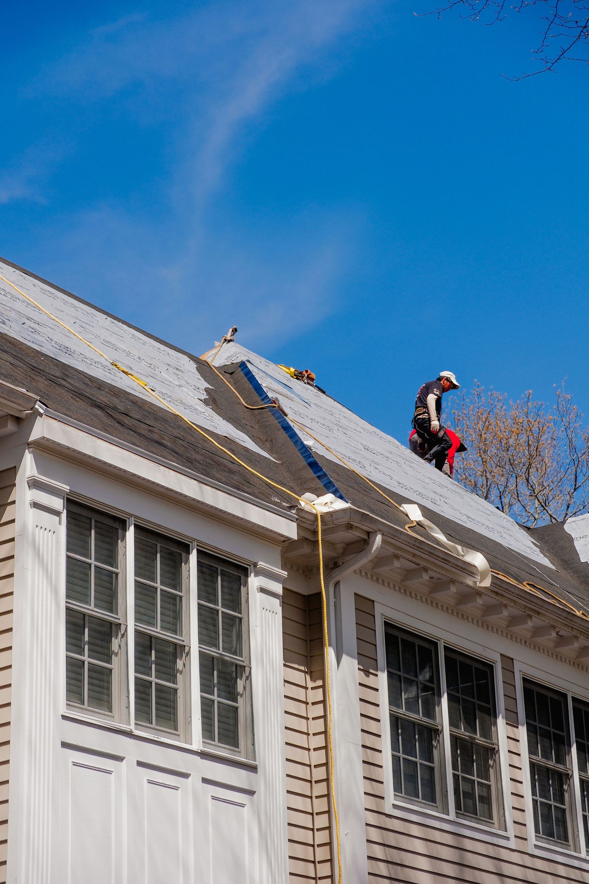 Person working on a roof with safety harness, a bright blue sky, and windows.