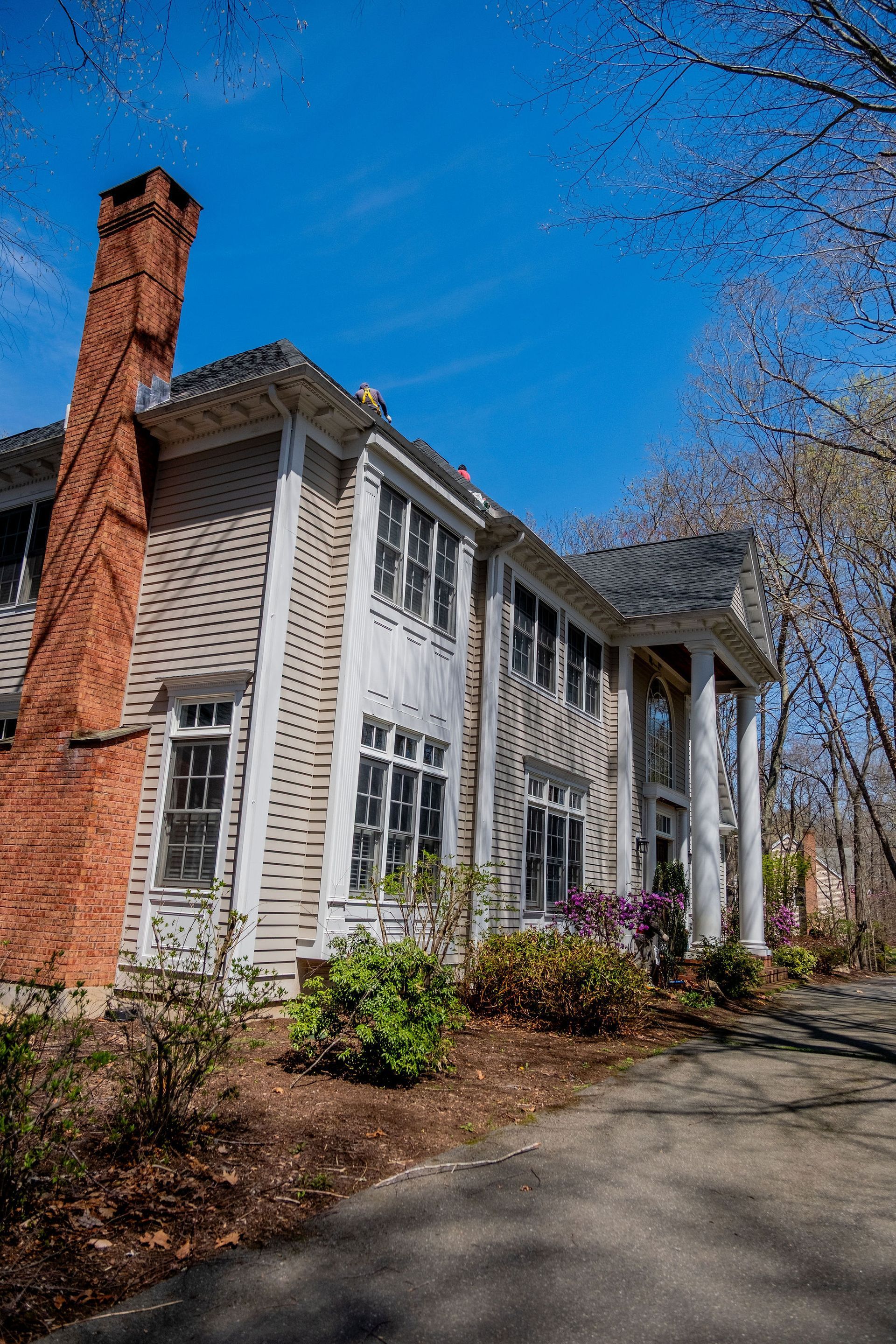 Two-story gray house with brick chimney, white columns, and landscaping under a blue sky.