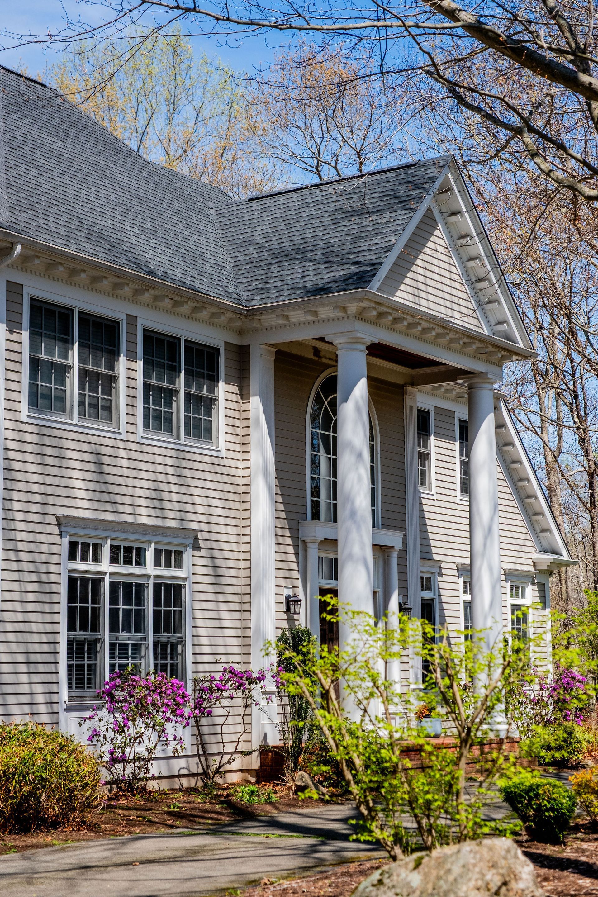 Beige house with white pillars, dark gray roof, and flowering bushes in the front yard.