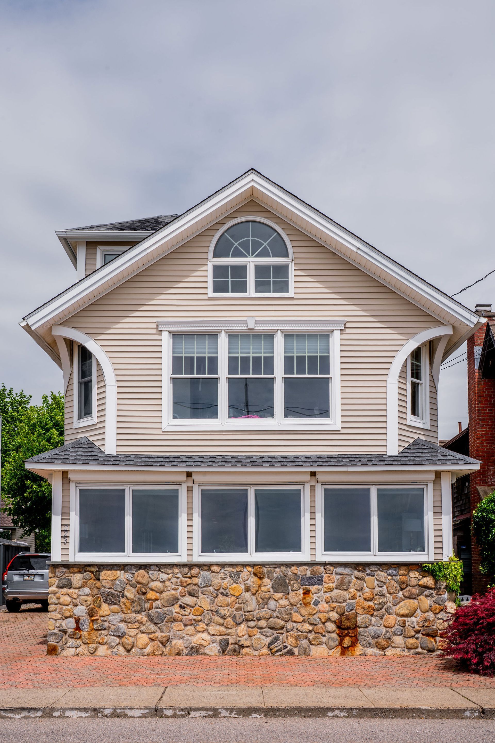 Two-story house with tan siding, stone base, arched window, and a small car parked outside.