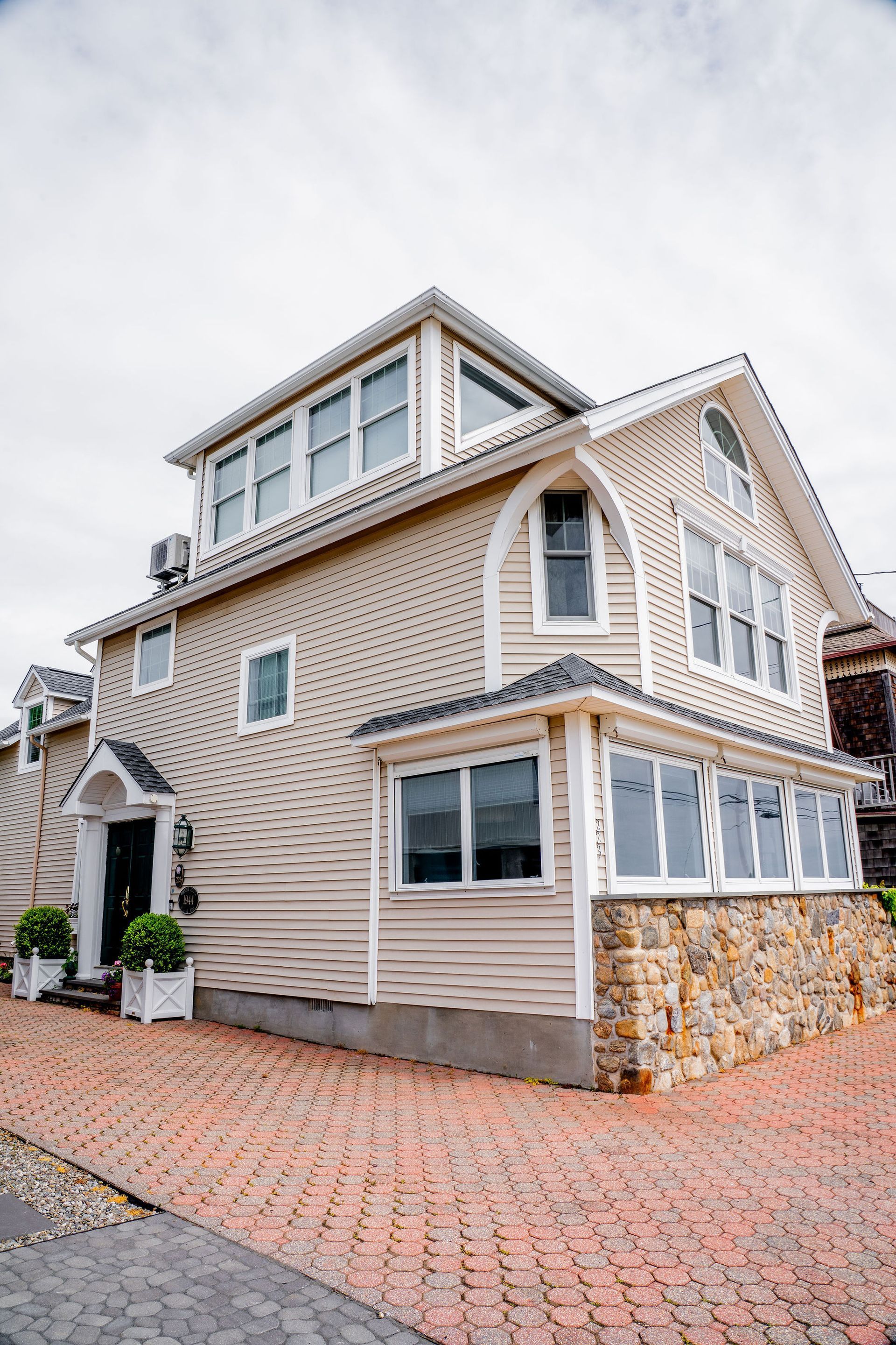 Beige two-story house with stone base and brick walkway under overcast sky.