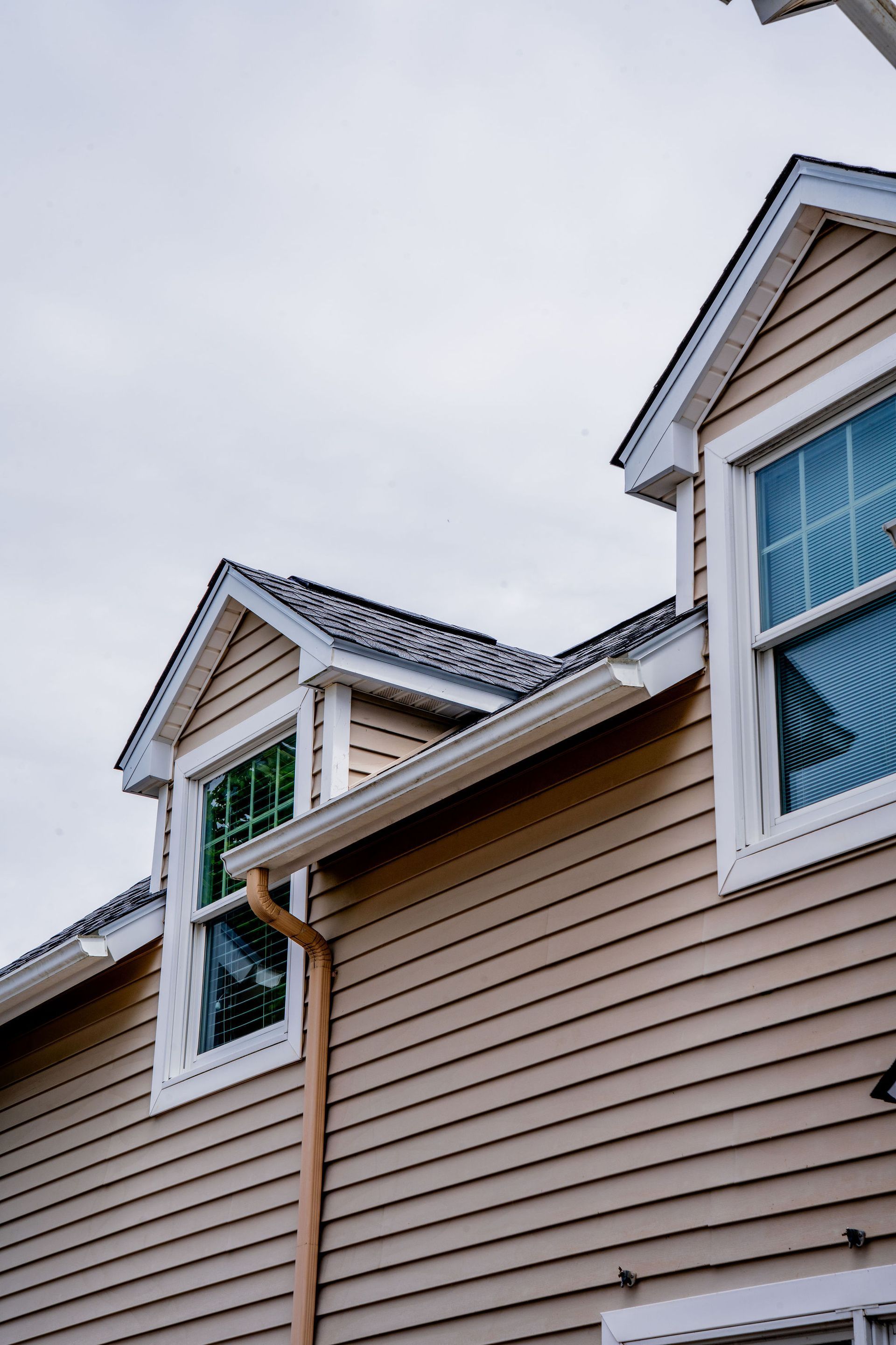 Tan house with two dormers, white-trimmed windows, and tan siding, under a gray sky.