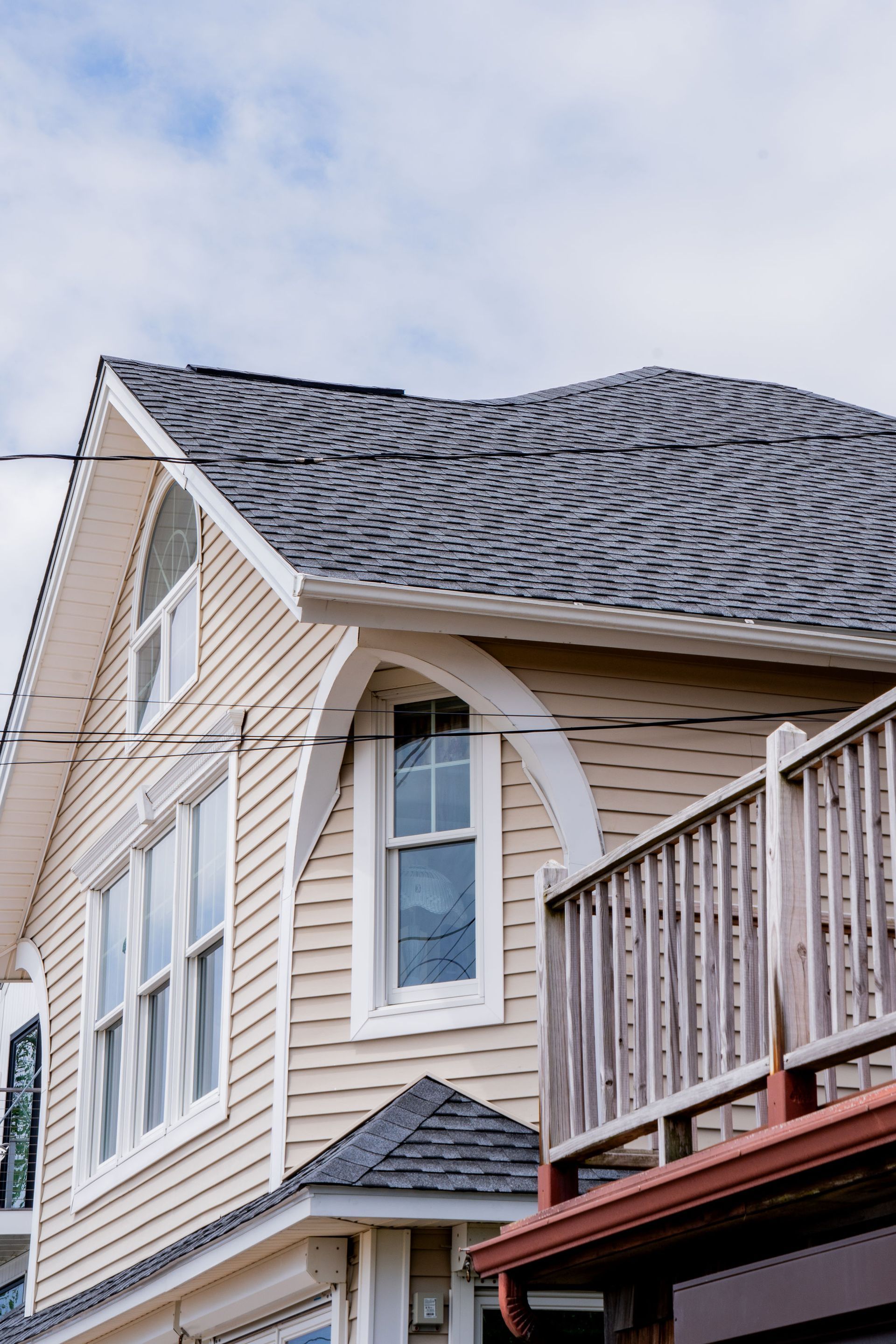 Beige house with gray shingle roof, arched windows, and wooden deck against a cloudy sky.