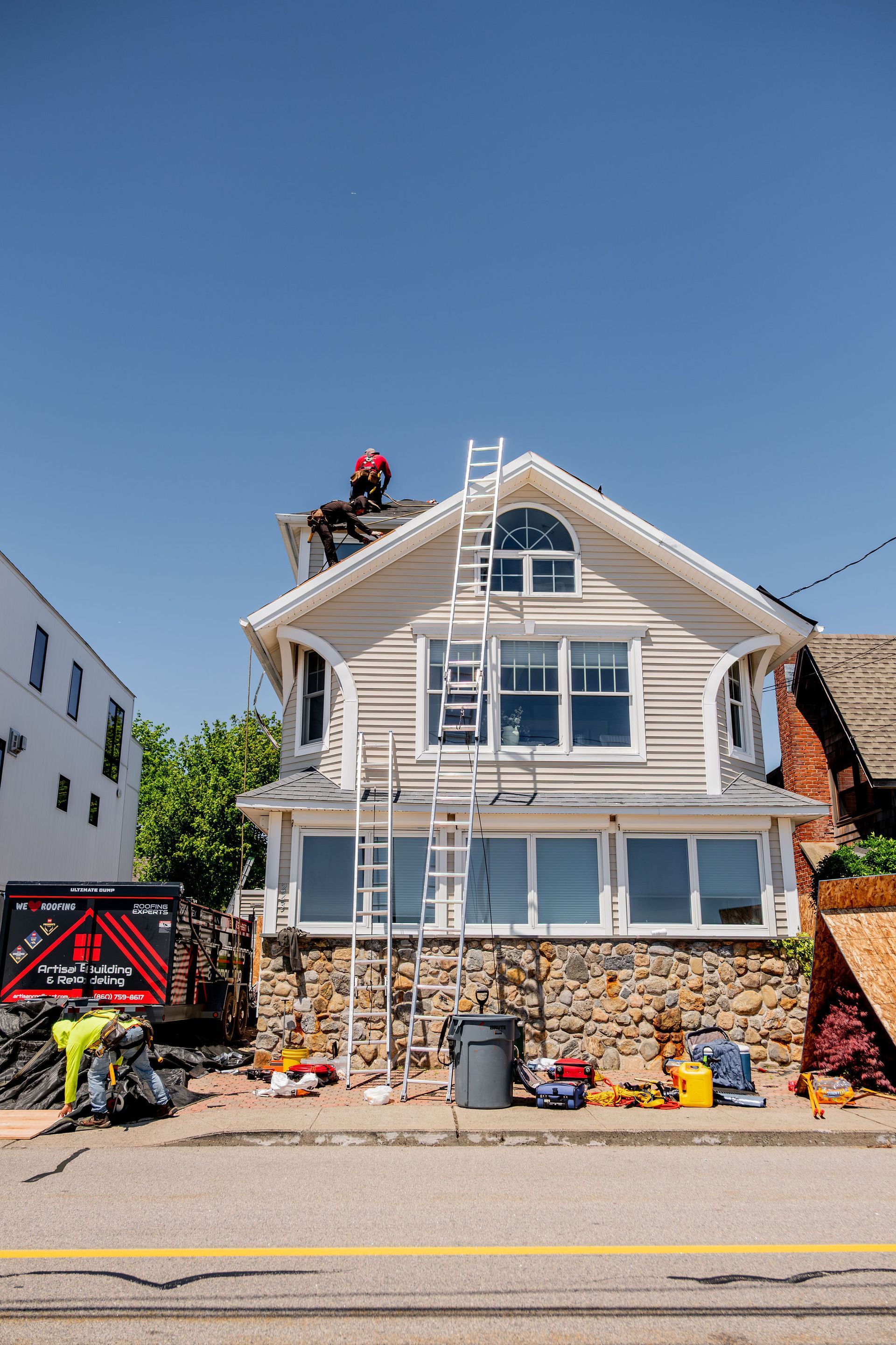 House under construction, workers on roof and ground. Ladder on the side. Clear blue sky.