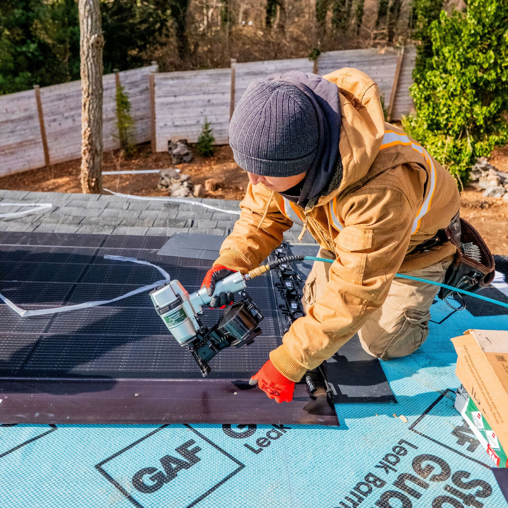 Roofer kneeling, using a nail gun to attach solar shingles to a roof. 