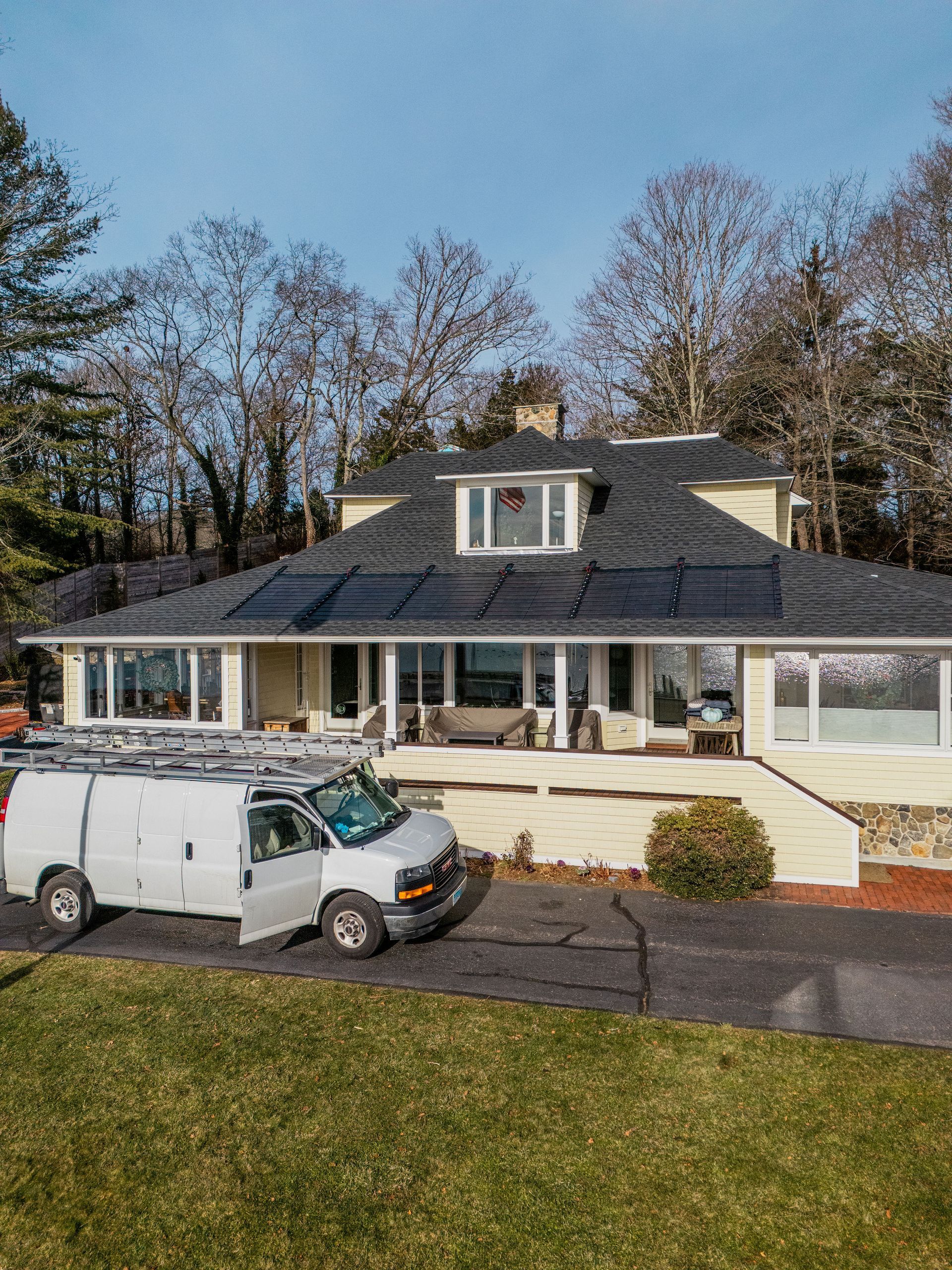 White van parked in front of a yellow house with a black roof and solar panels. Trees in the background.