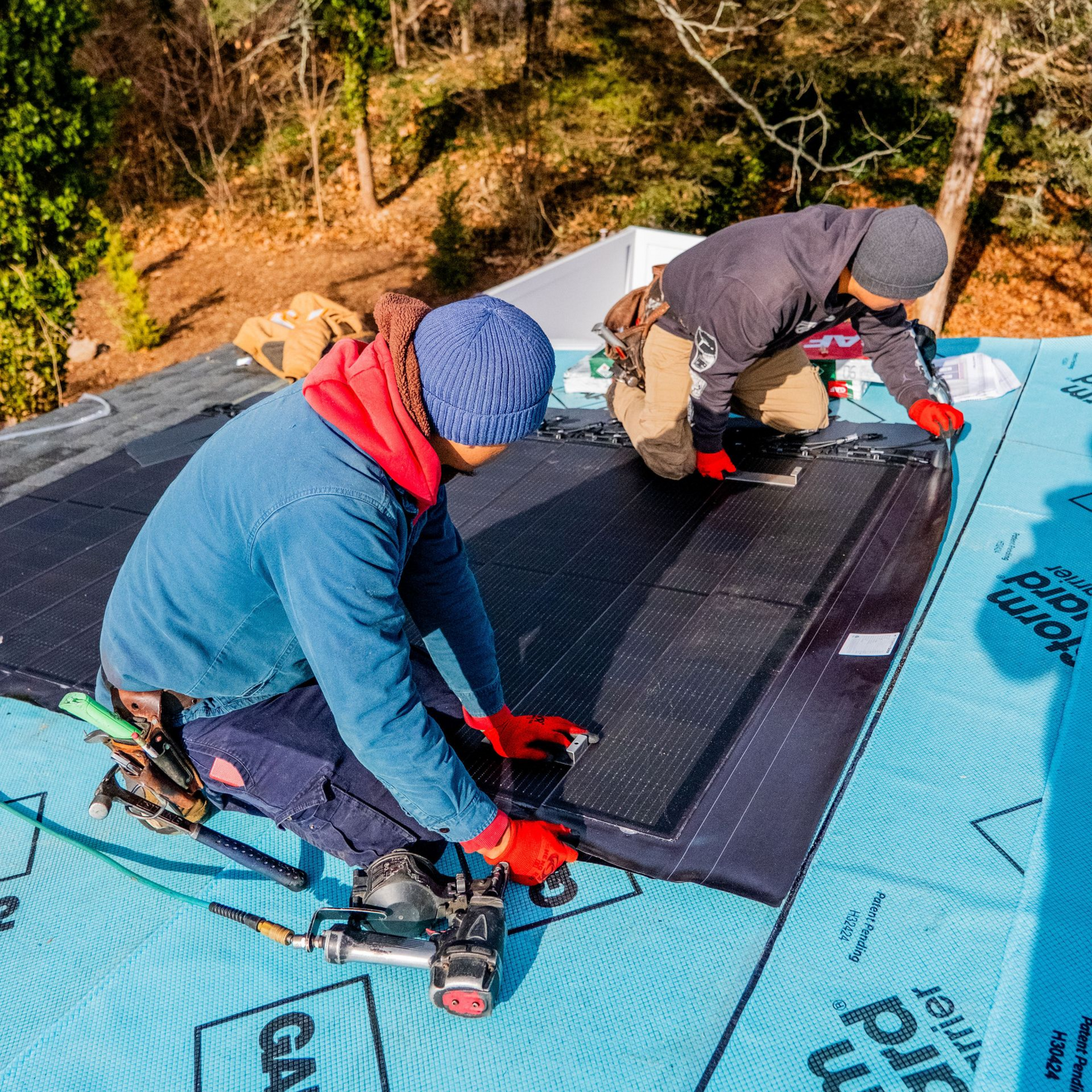 Two workers installing solar panels on a rooftop. Blue tarp and panels. One using a nail gun.