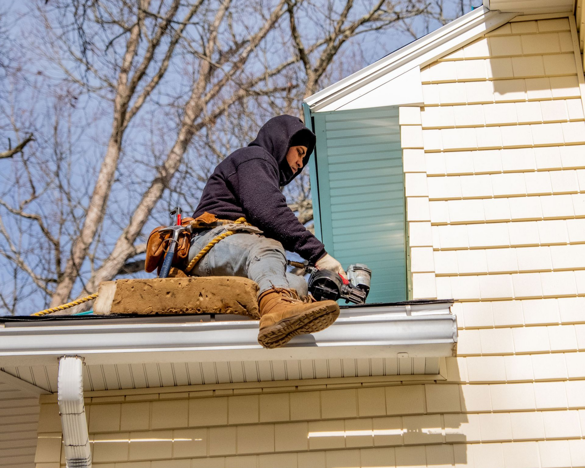 Roofer seated on a roof, installing siding on a light yellow house, using a nail gun.
