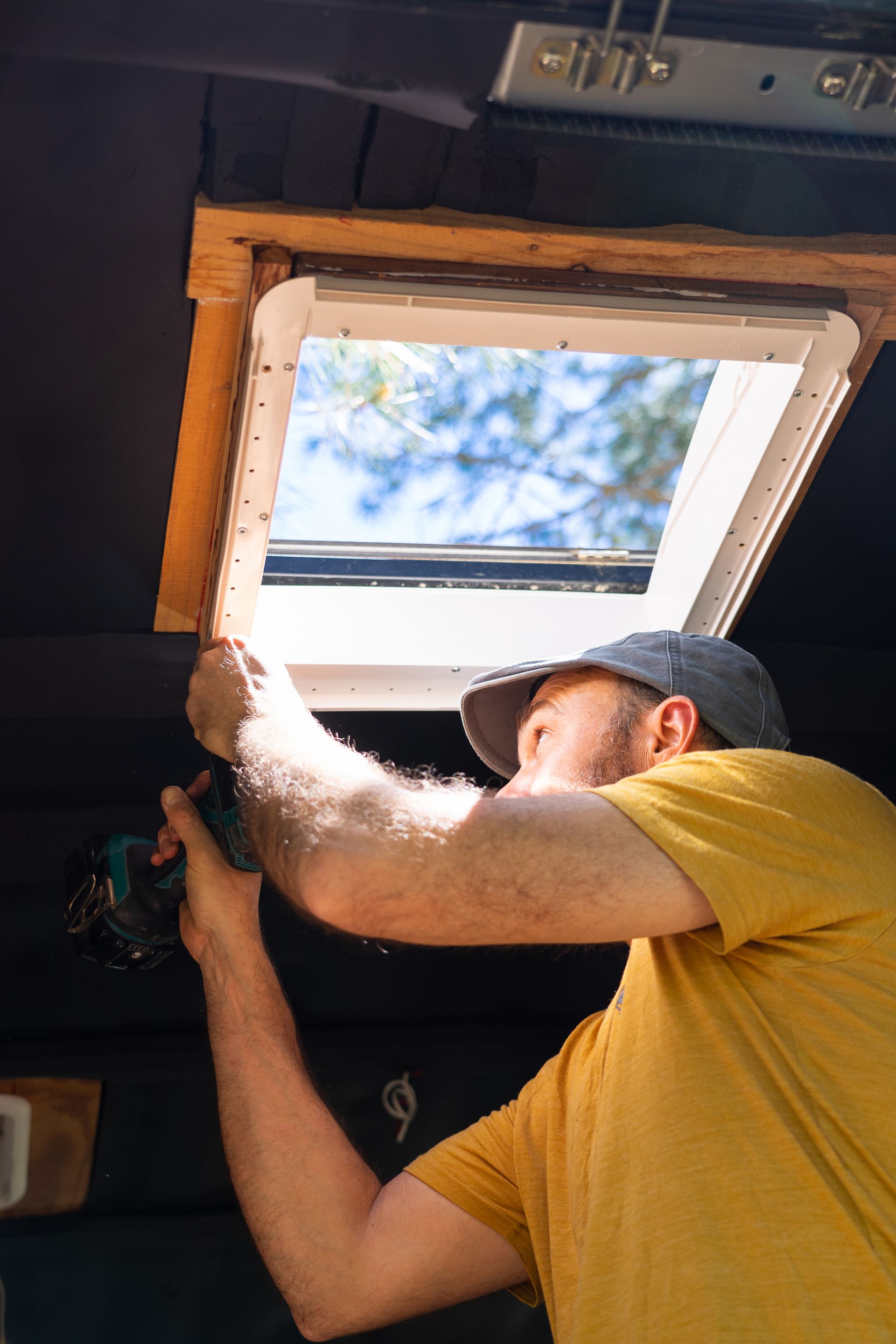 A person using a drill to install a skylight in a dark-colored interior. Sunlight streams through the opening.
