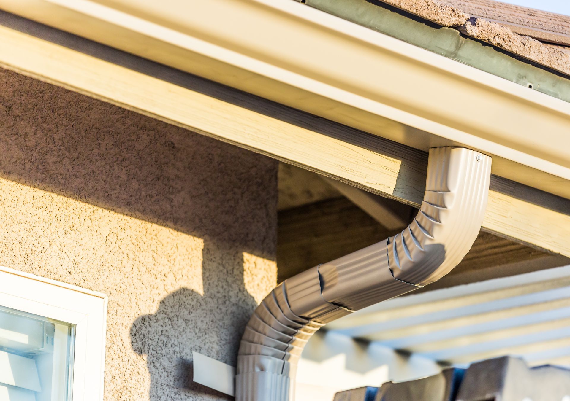 Gutter downspout curving from a roof corner beside a stucco wall and window