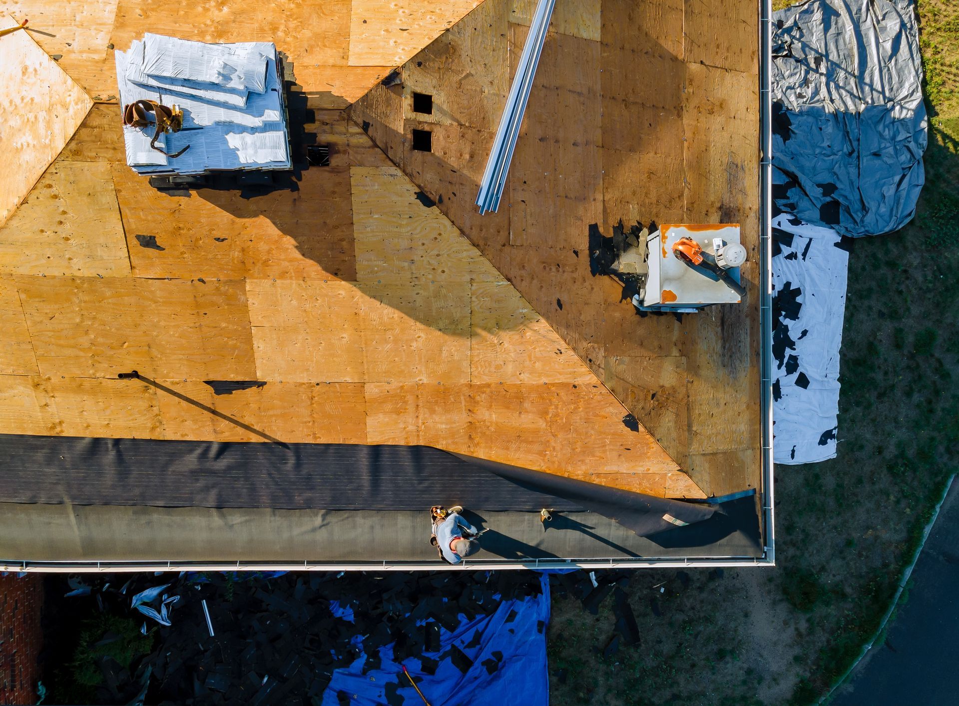 An overhead view of a roof under construction, showing plywood sheathing, roofing materials, and a worker applying underlay.