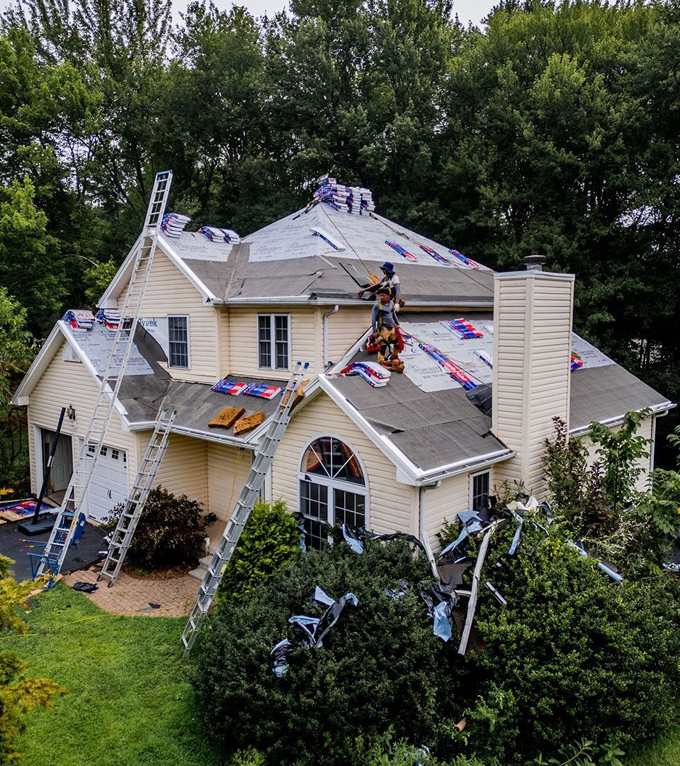Roofers working on a beige house, covered with protective material. Ladders leaned against the roof.