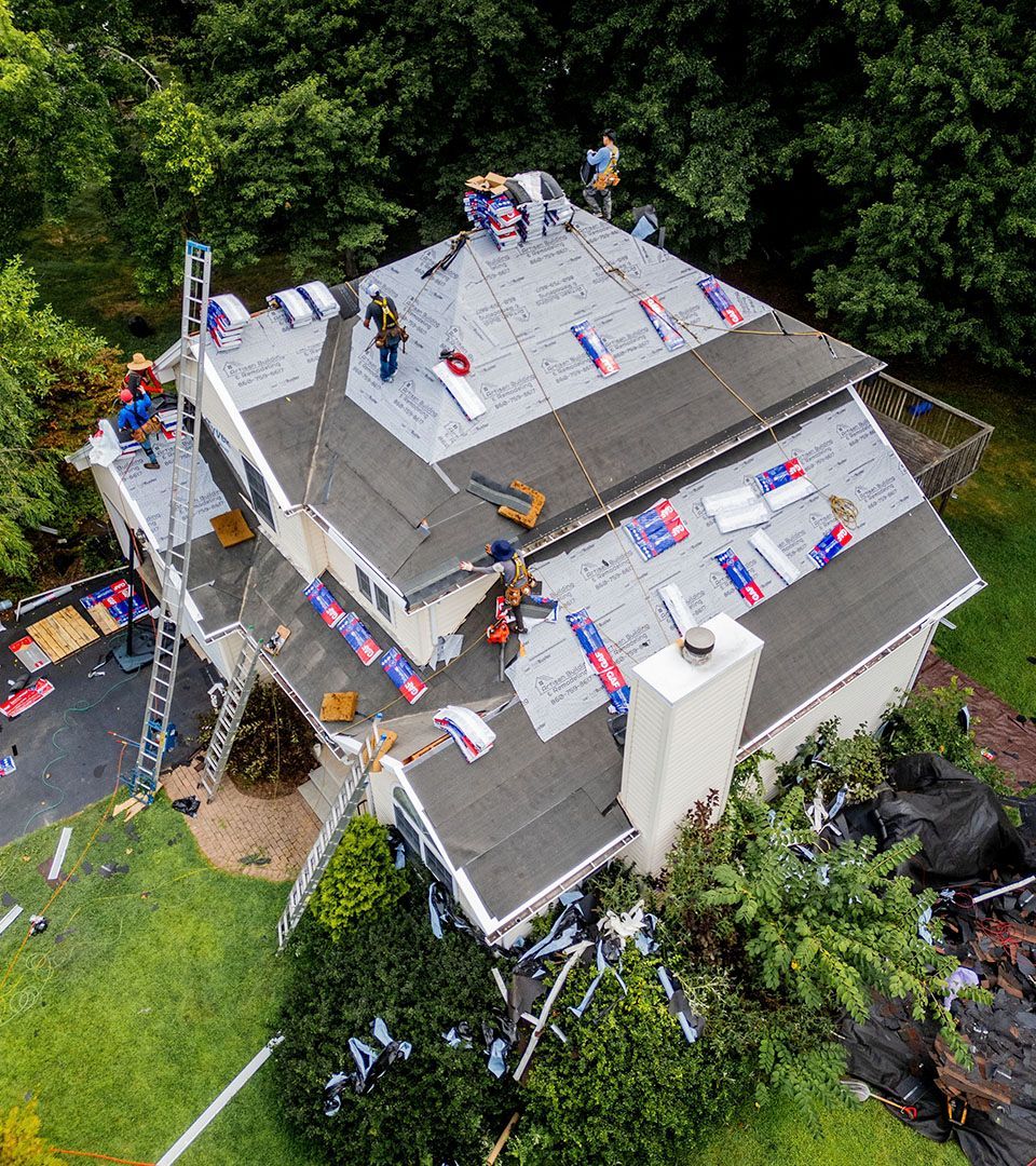 Roofers working on a house roof, laying shingles. Green trees surround the house.