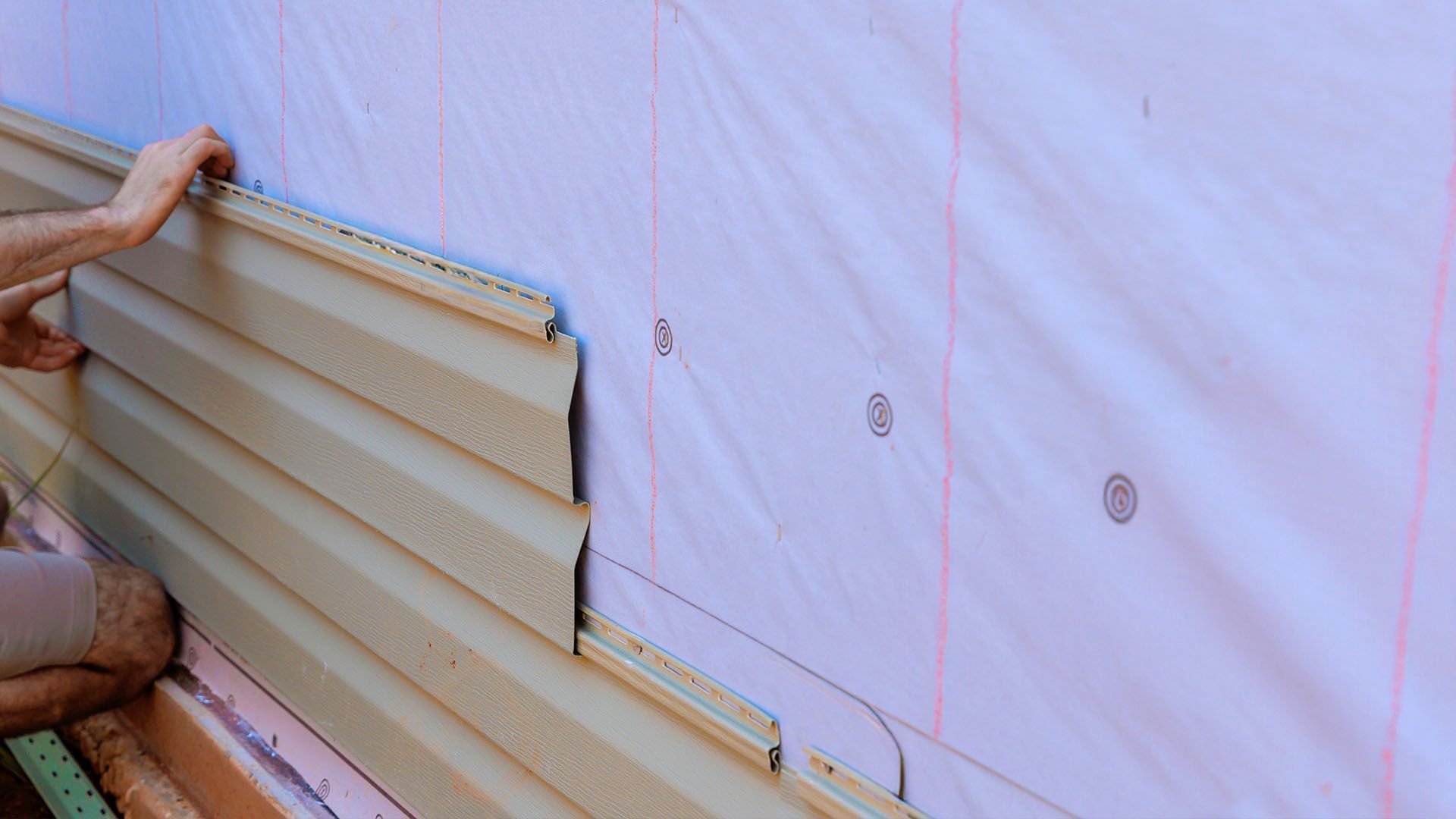 Person installing tan vinyl siding on a pink-sheathed wall.