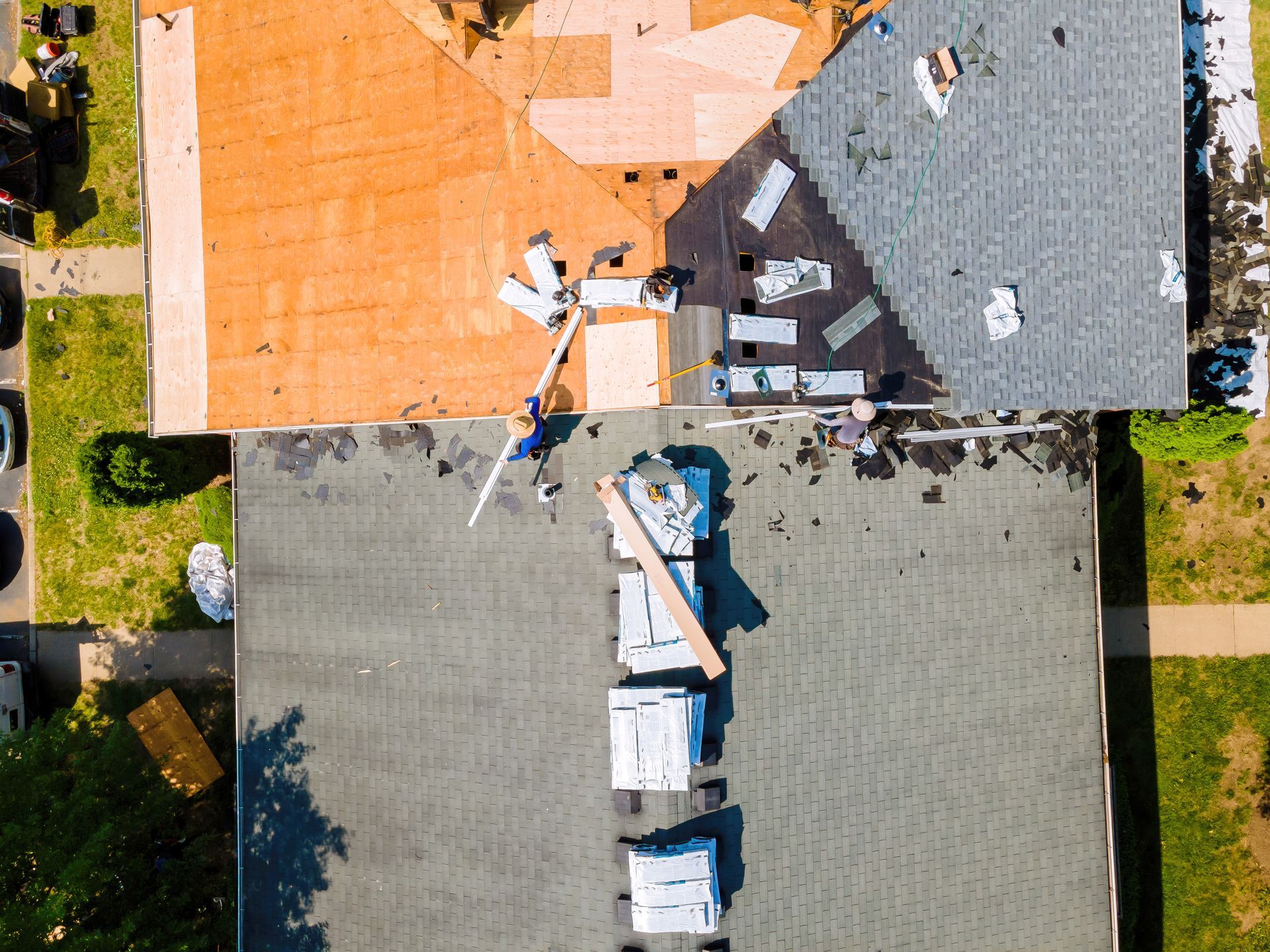 Overhead view of a building roof under construction: orange, gray and green sections; workers are present, surrounding grassy areas.