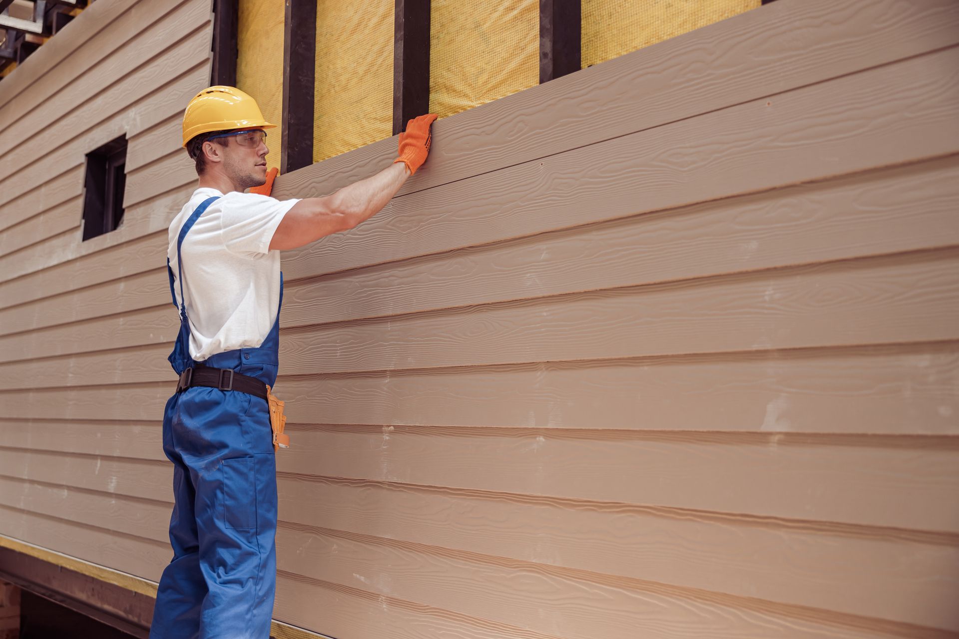 Worker in hard hat and gloves measuring beige siding on a house exterior