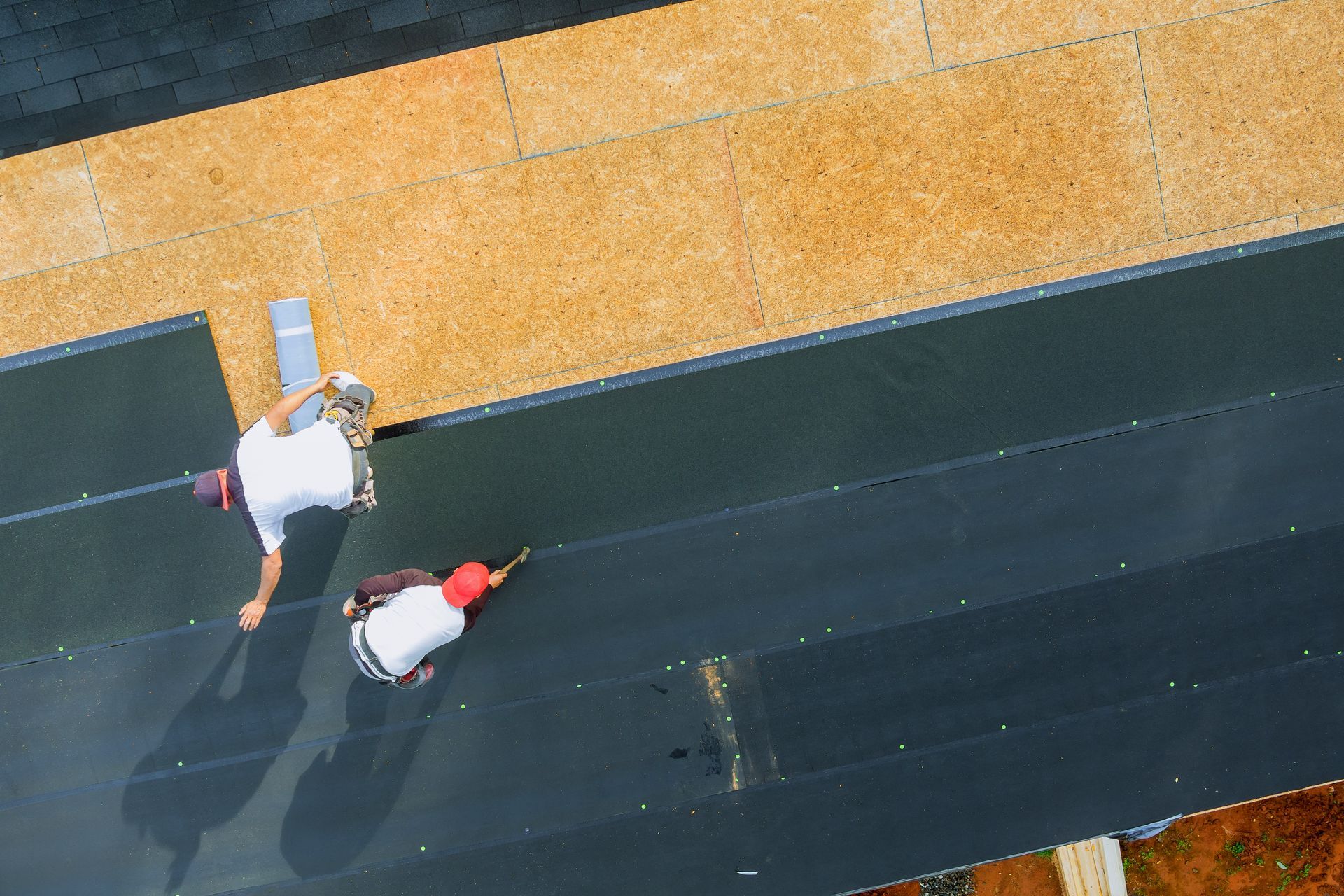 Two roofers on a black flat roof install roofing material, overhead view.