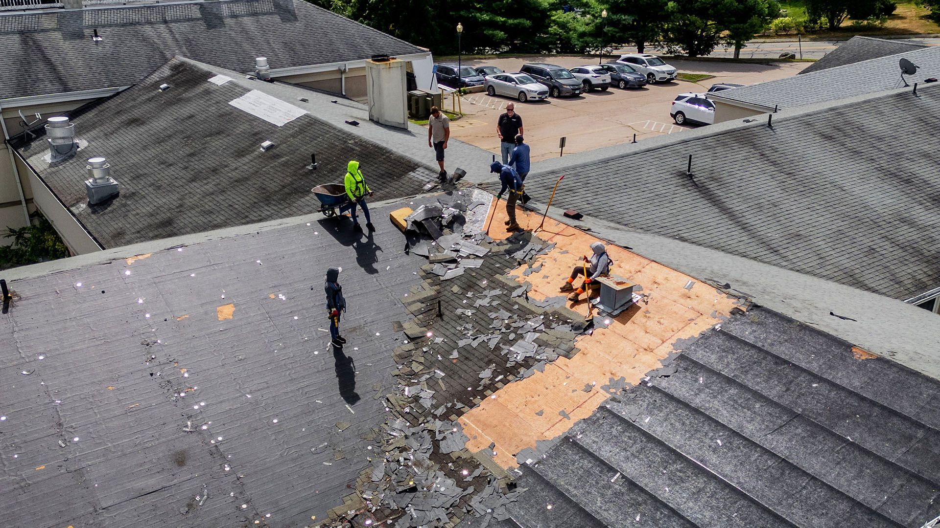 Workers repairing a damaged roof. Gray shingles, orange underlayment visible. Cars parked nearby.
