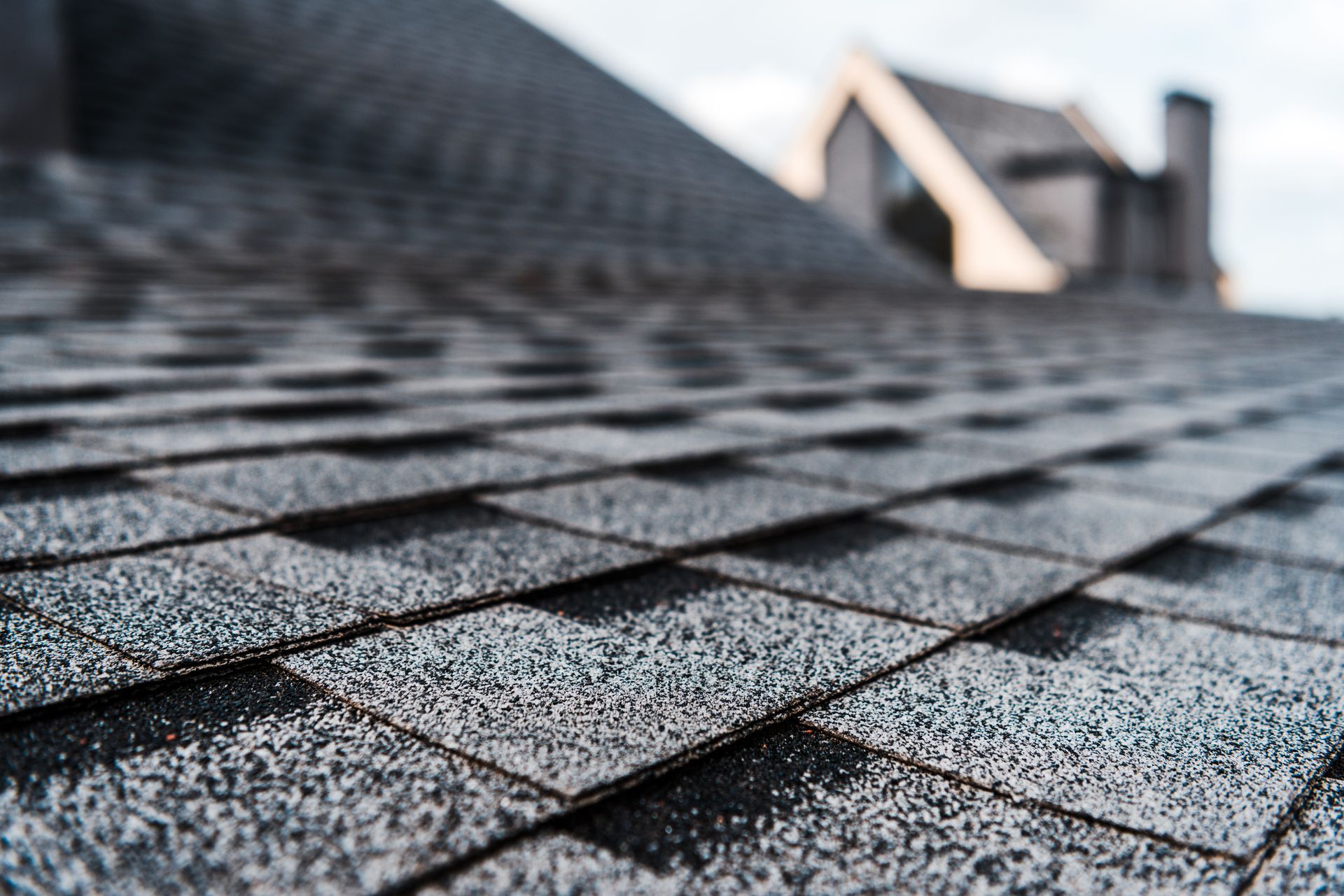 Close-up of gray asphalt roof shingles with blurred roofline and chimney in the background