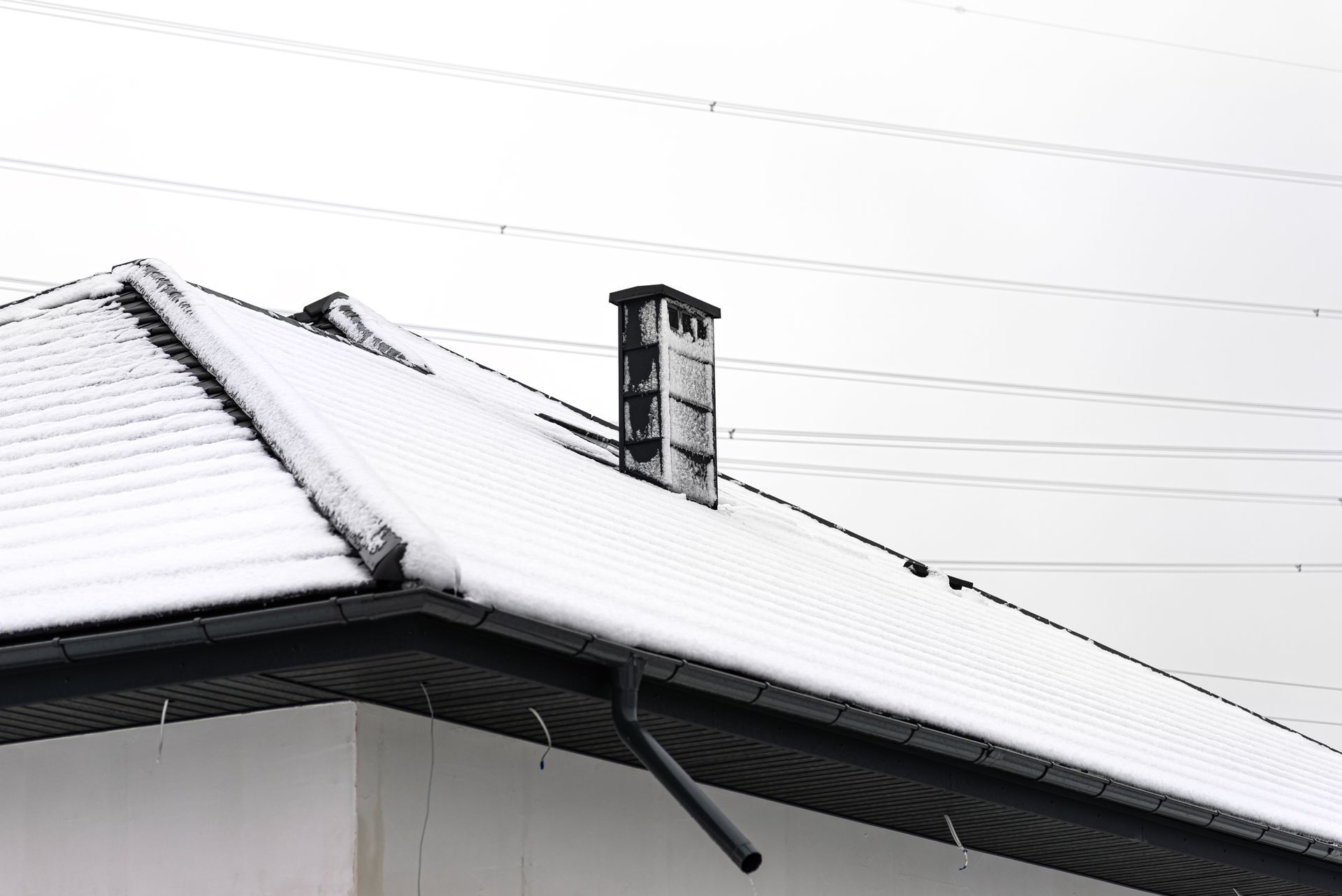 Snow-covered roof of a building with a chimney and a black gutter; power lines in the background.