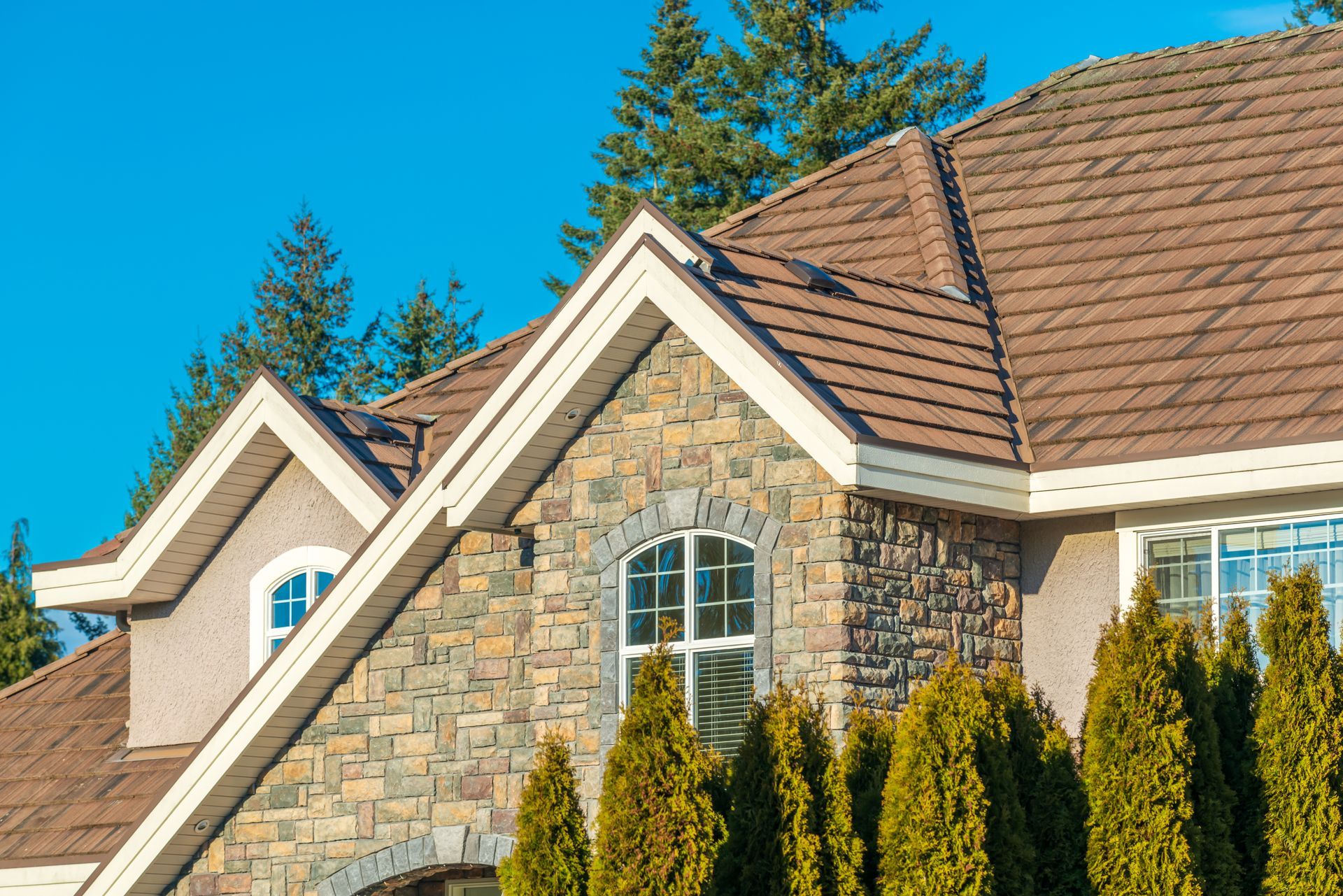 Stone-fronted house with brown roof and two gabled dormers. Tall green trees and blue sky.
