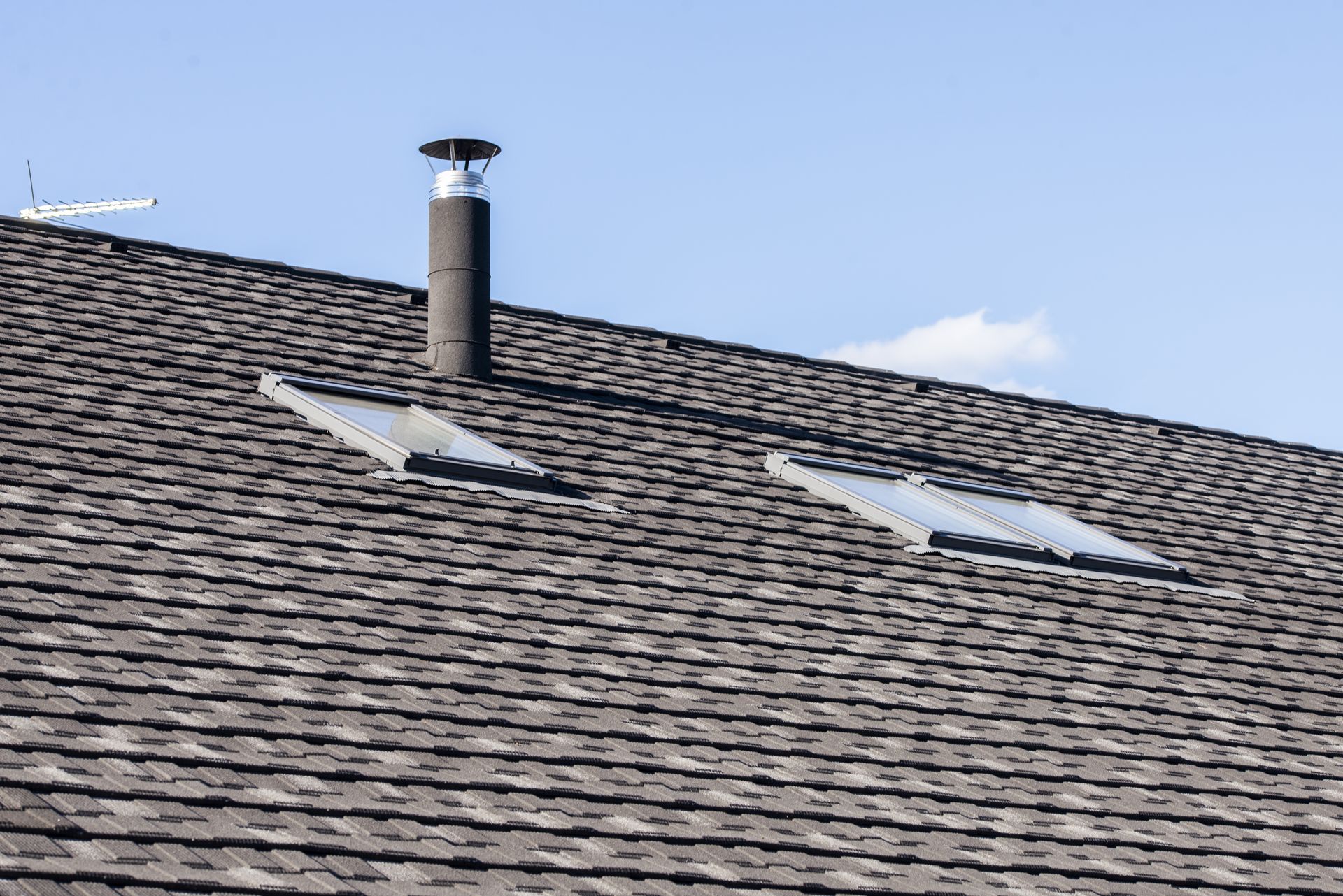 Dark shingled roof with two skylights and a chimney against a blue sky.