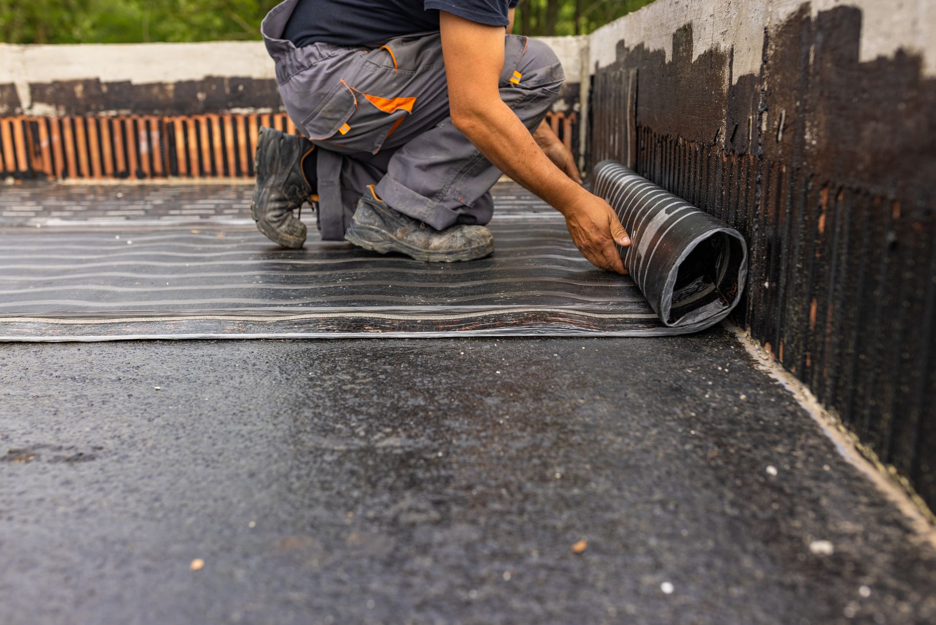 Person installing waterproofing membrane on a construction site. Black material against a black tar surface.