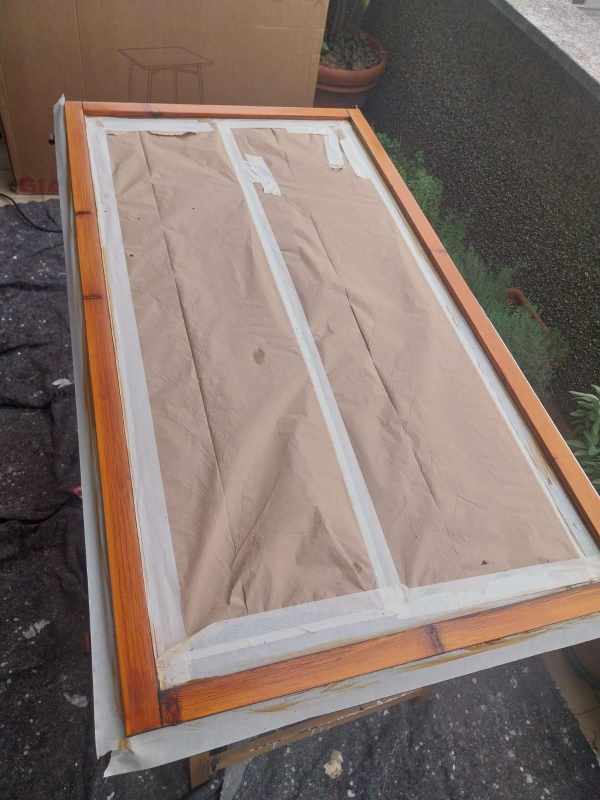 Opera d'arte incorniciata in legno, ricoperta di carta marrone e nastrata. Esposta all'aperto, su una superficie nera.