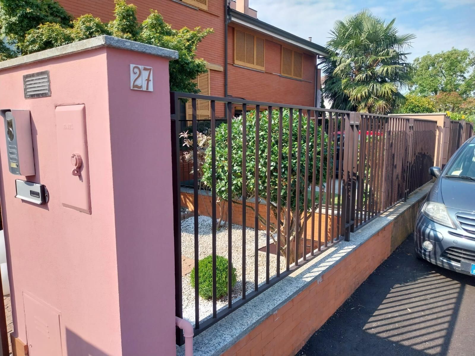 Recinzione in mattoni rosa con sbarre di ferro, contenente arbusti e pietre. Edificio sullo sfondo. Auto parcheggiata sulla destra.