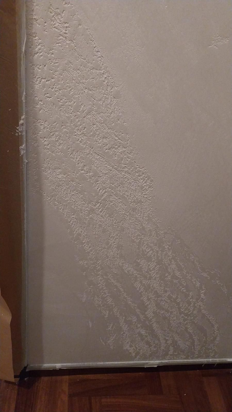 Parete bianca con danni causati dall'acqua che scendono verso il basso; battiscopa e pavimento in legno visibili.