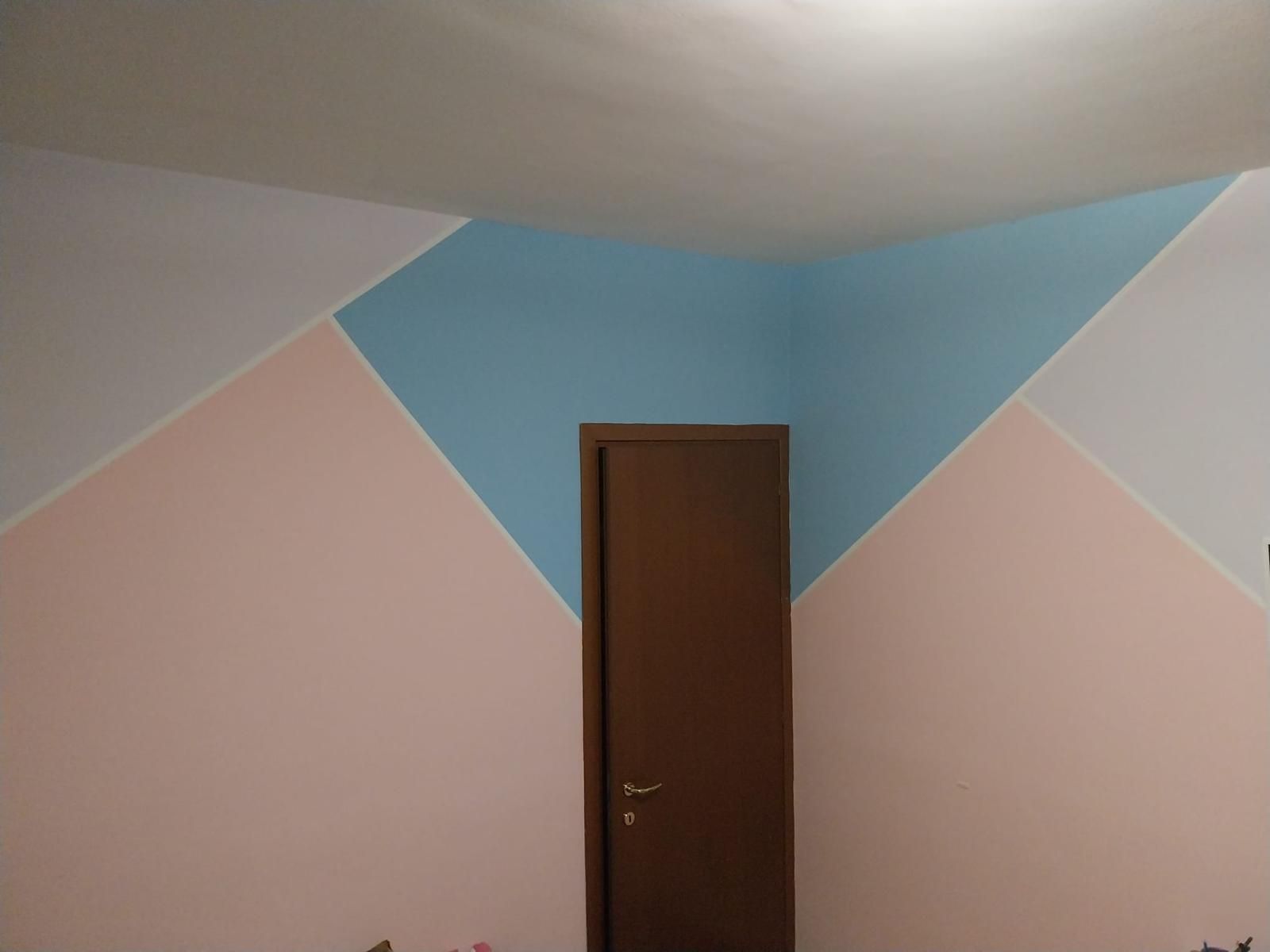 Interno di una stanza con pareti dipinte con motivi geometrici: triangoli blu, rosa e bianchi che circondano una porta marrone.
