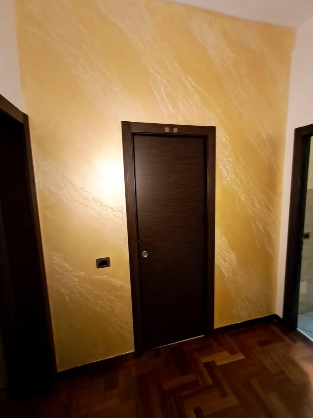 Porta scura in corridoio dai toni dorati. Pareti con finitura ruvida. Pavimento in legno marrone.
