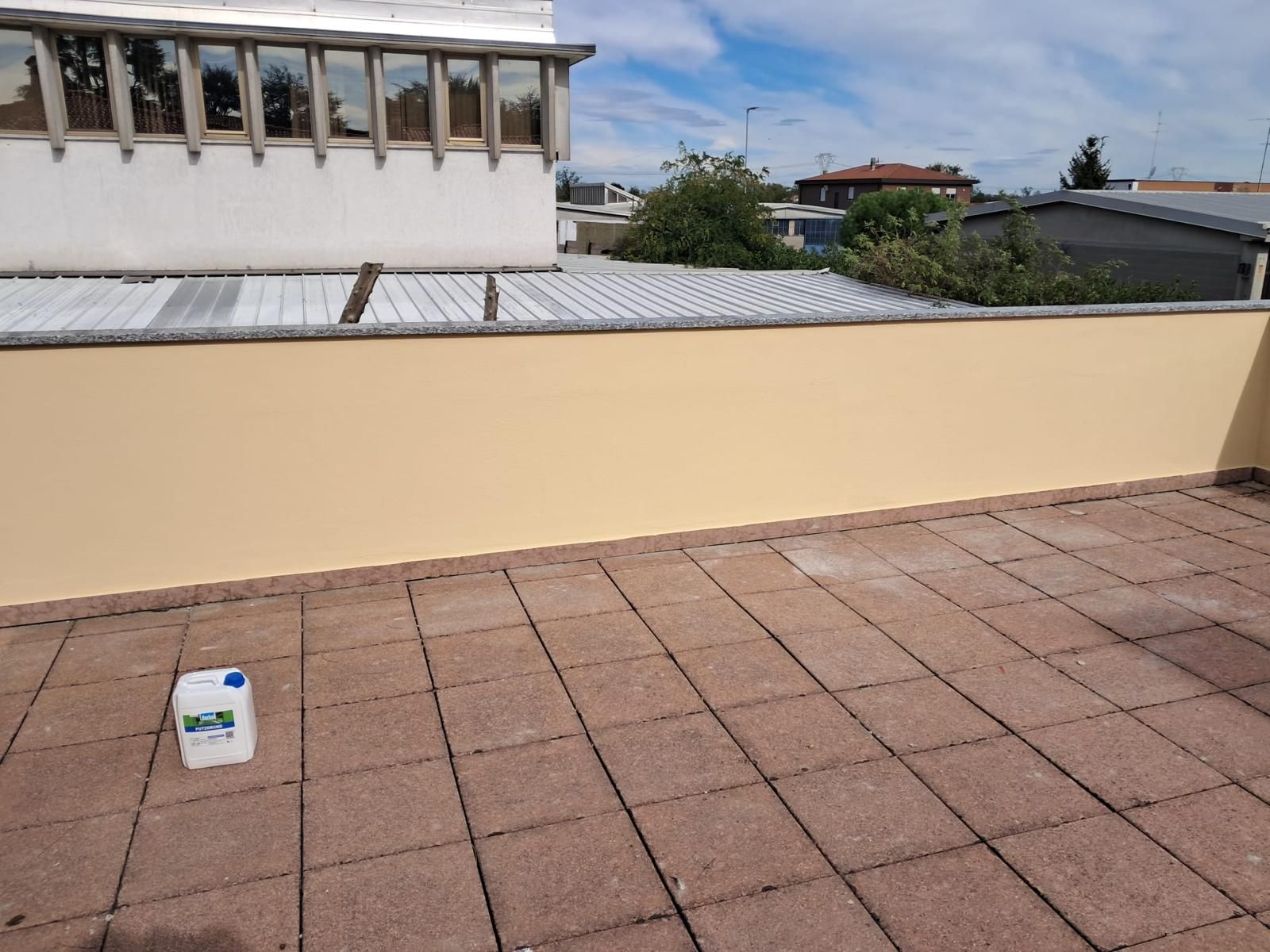 Tetto con pavimentazione in mattoni, muro giallo e un edificio sullo sfondo. Un container bianco è appoggiato sulla pavimentazione.