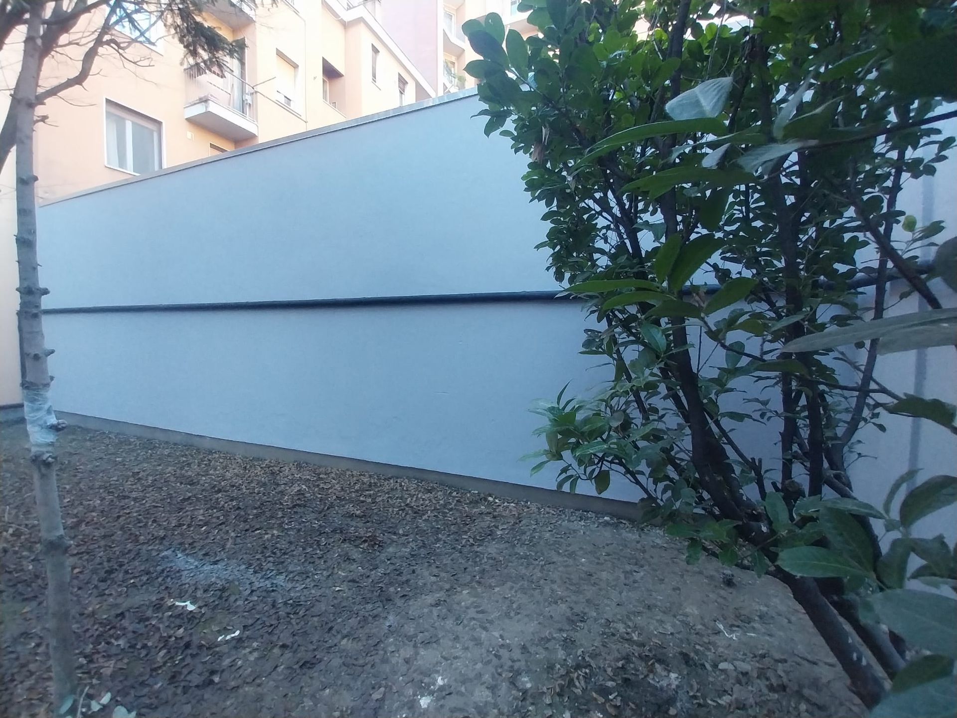 Muro grigio con una linea orizzontale scura, terreno ghiaioso e vegetazione di fronte. Edificio visibile dietro il muro.