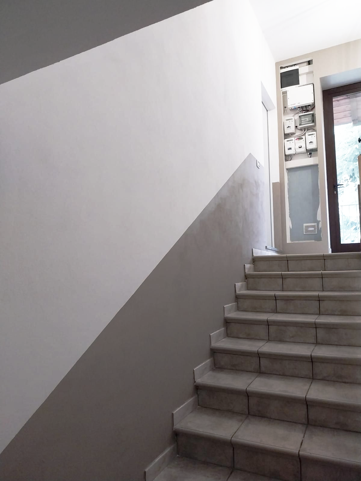 Scala in un edificio con pareti dipinte di grigio e bianco, che conduce a una porta con contatori elettrici sul muro.